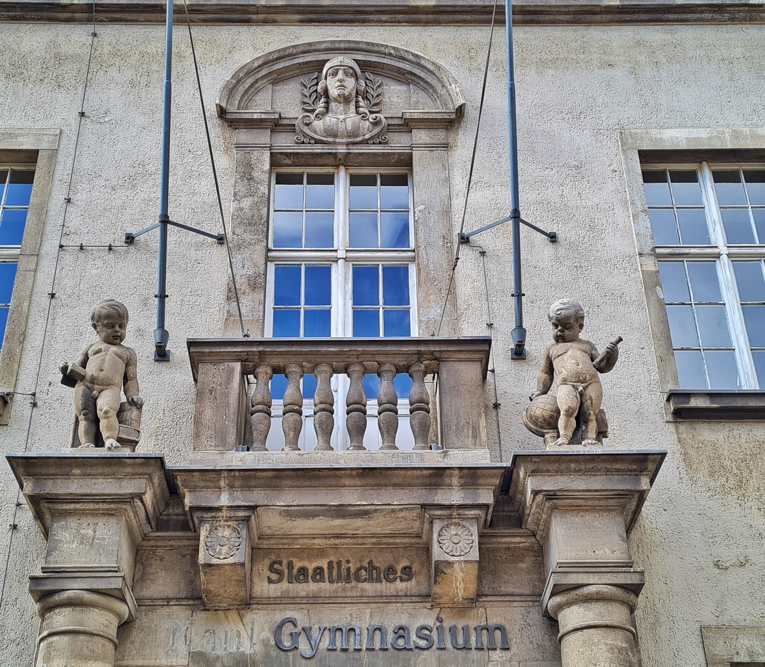 Ein Teil einer Gebäudefassade mit einem Balkon. Darunter steht "Staatliches Gymnasium". Hinter dem Balkon ist ein Fenster. Darüber der Kopf eines jungen Menschen. Links und rechts vom Balkon jeweils Puttenfiguren.