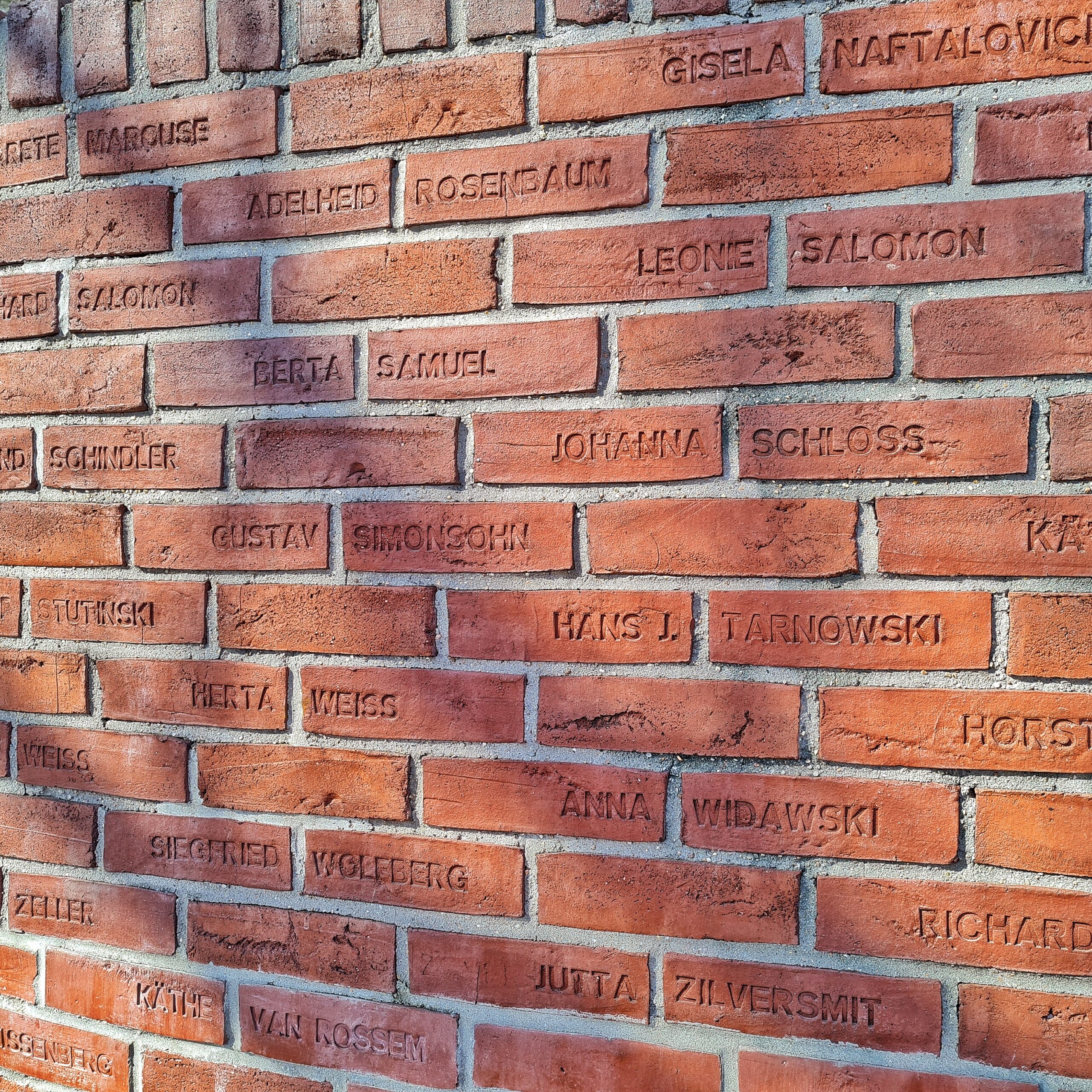 Eine Backsteinmauer. In die Steine sind Vornamen und Nachnamen eingeritzt.
