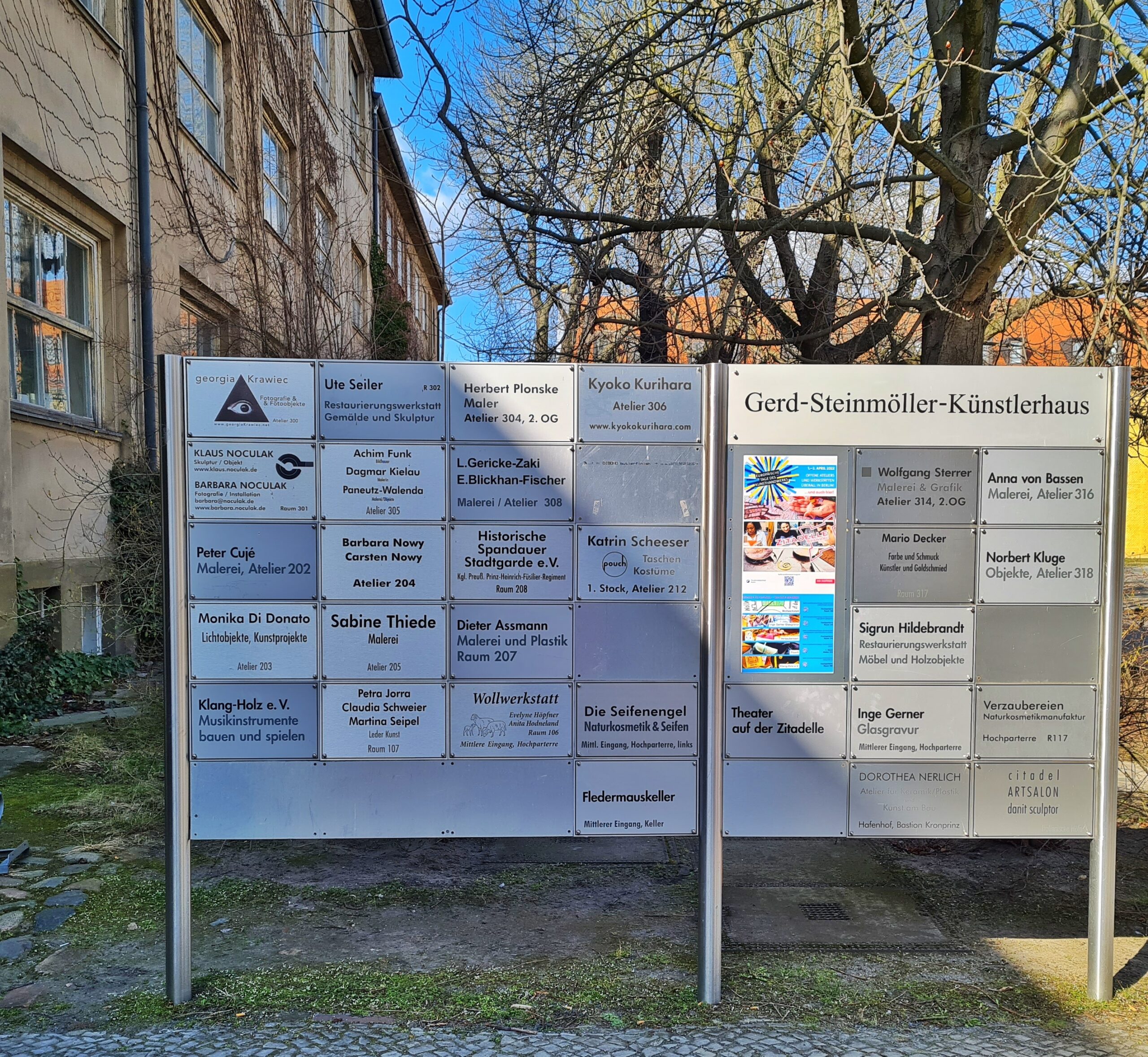 Links ist ein Gebäude erkennbar. Frontal zur Kamera steht ein großer Metallaufsteller. Darauf steht "Gerd-Steinmöller-Künstlerhaus". Außerdem sind darauf ganz viele kleine Schilder mit dem Hinweis auf Ateliers und Künstlerinnen und Künstler angebracht.