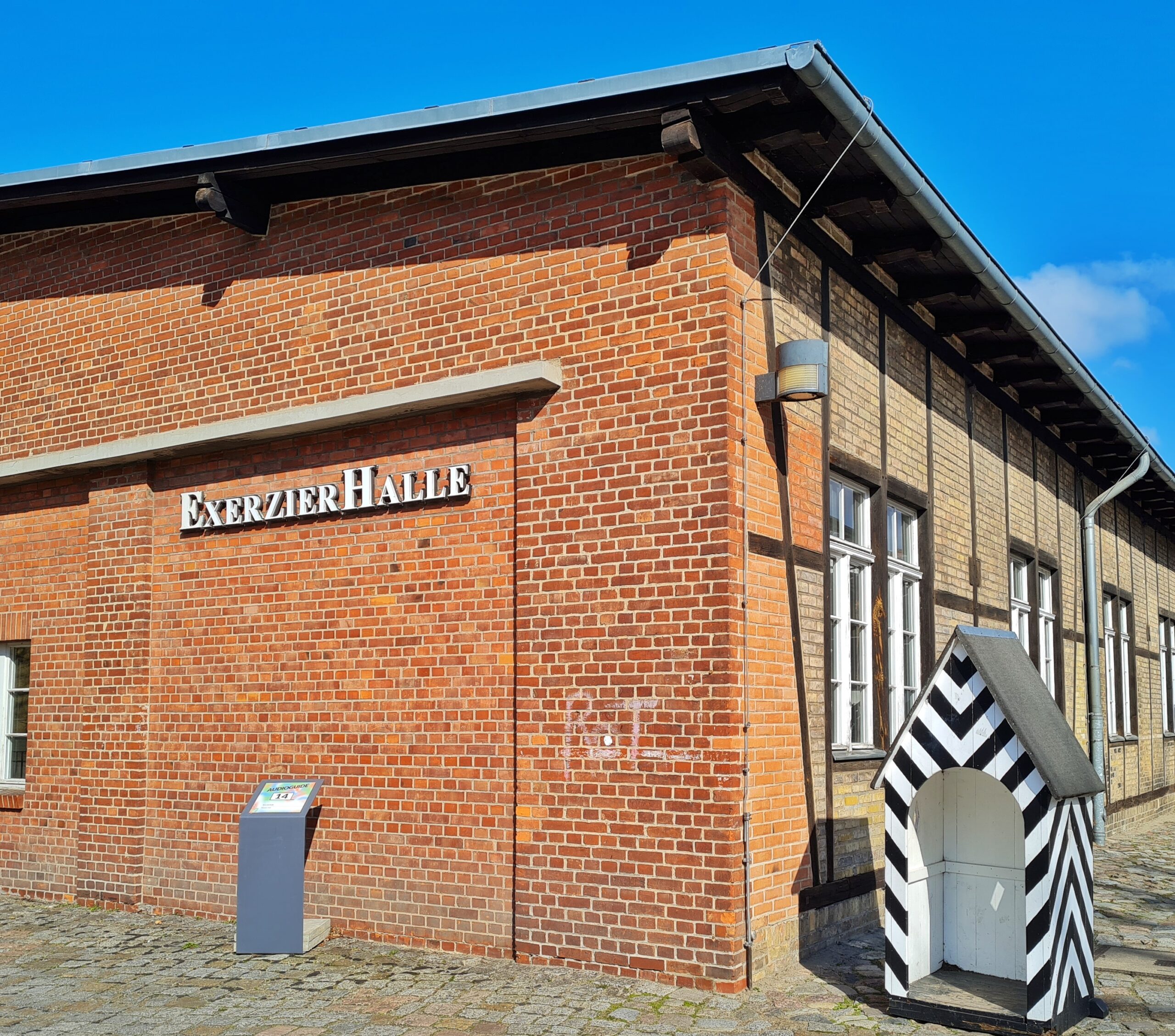 Ein Gebäude mit der Aufschrift "Exerzierhalle". Rechts davon ein Unterstand für einen Wachposten (weiß mit schwarzen Streifen), allerdings sehr klein.