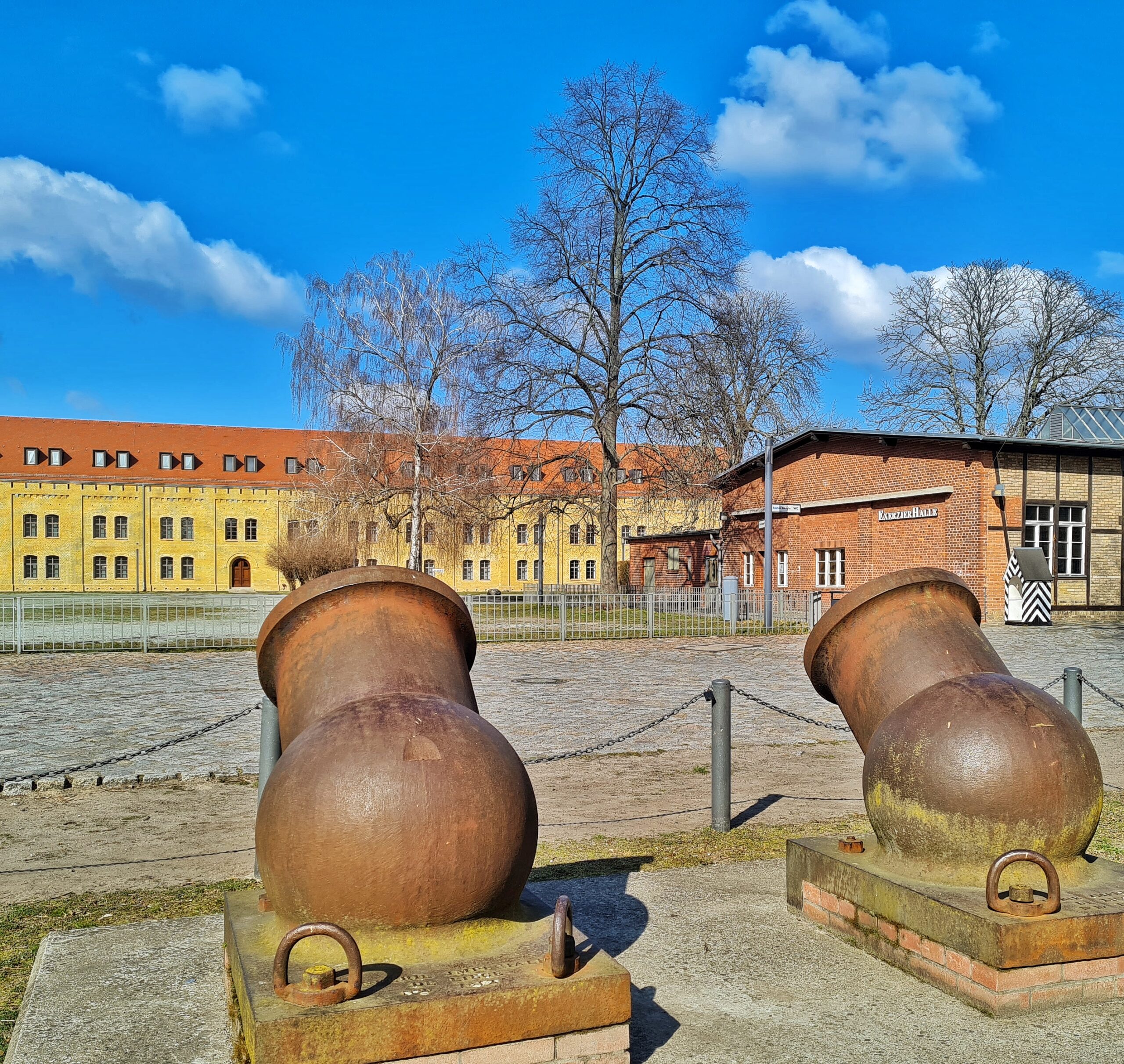 Im Hintergrund wieder das langgestreckte, gelbe Gebäude. Rechts ein weiteres Gebäude und drei kahle Bäume. Im Vordergrund zwei Kanonen.