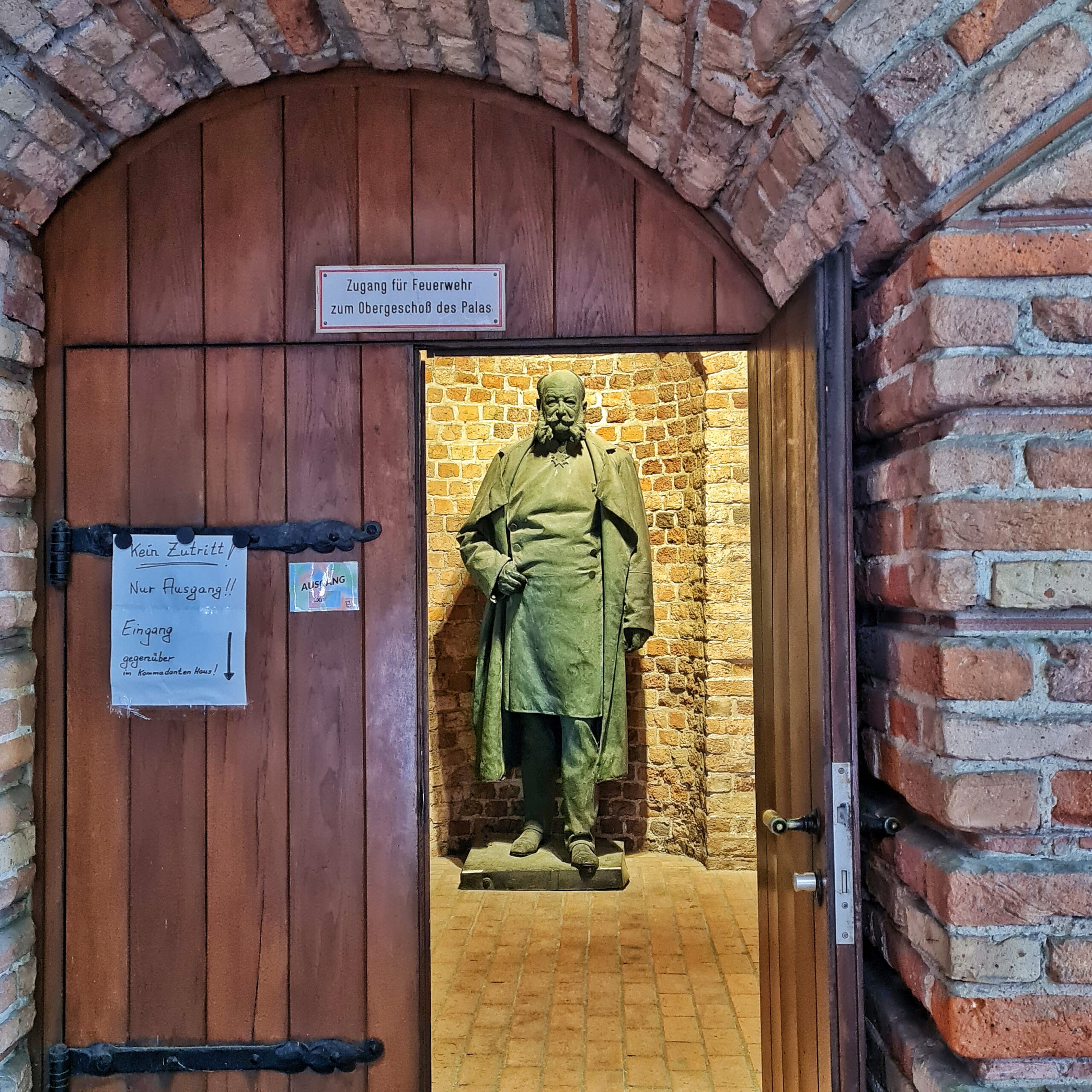 Blick durch eine halboffene Rundbogentür in einen gemauerten Raum, in dem die Statie eines alten, bärtigen Mannes steht. Auf der Tür ein Schuld "Zugang für Feuerwehr zum Obergeschoß des Palas" und ein Zettel "Kein Zutritt! Nur Ausgang!! Eingang gegenüber"