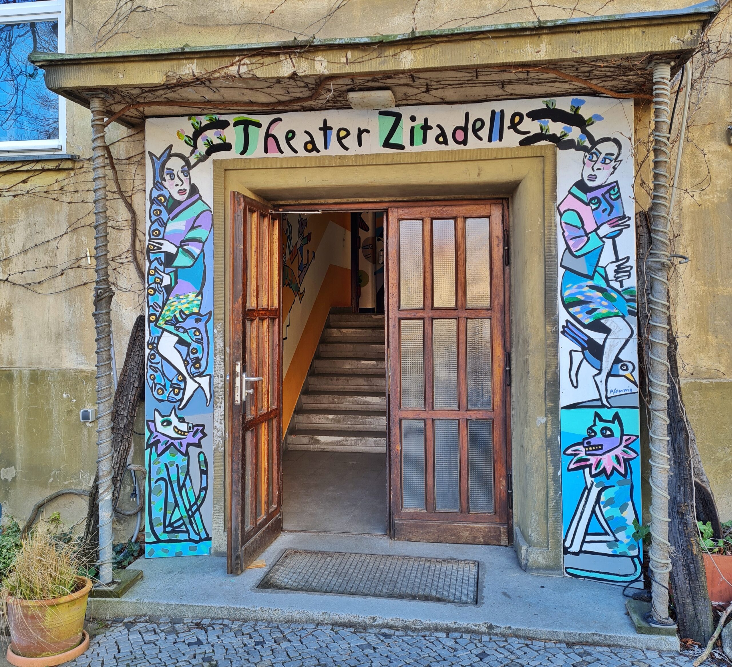 Der Eingang eines Gebäudes. Eine Hälfte der Sprossentür steht offen und zeigt innen Treppenstufen. Über dem Eingang steht in bunten Buchstaben "Theater Zitadelle" und die beiden Seiten des Eingangs sind mit Menschen und Tieren bemalt.