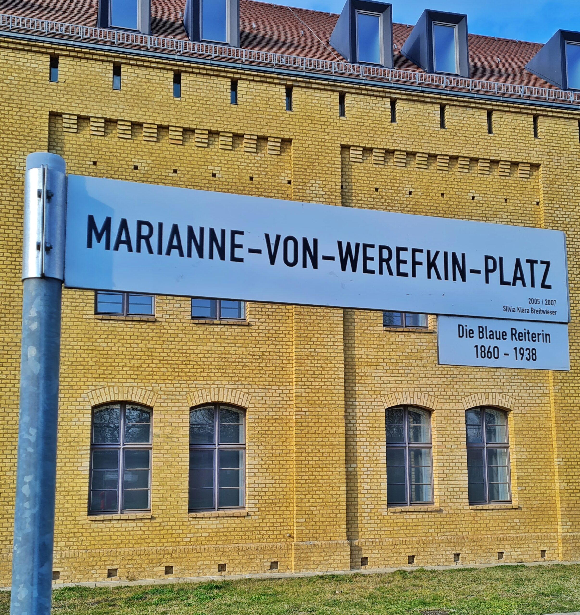 Im Hintergrund ein gelbes Backsteingebäude. Im Vordergrund ein Straßenschild "Marianne-von-Werefkin-Platz" mit einem kleinen Zusatzschild "Die blaue Reiterin 1860 - 1938".