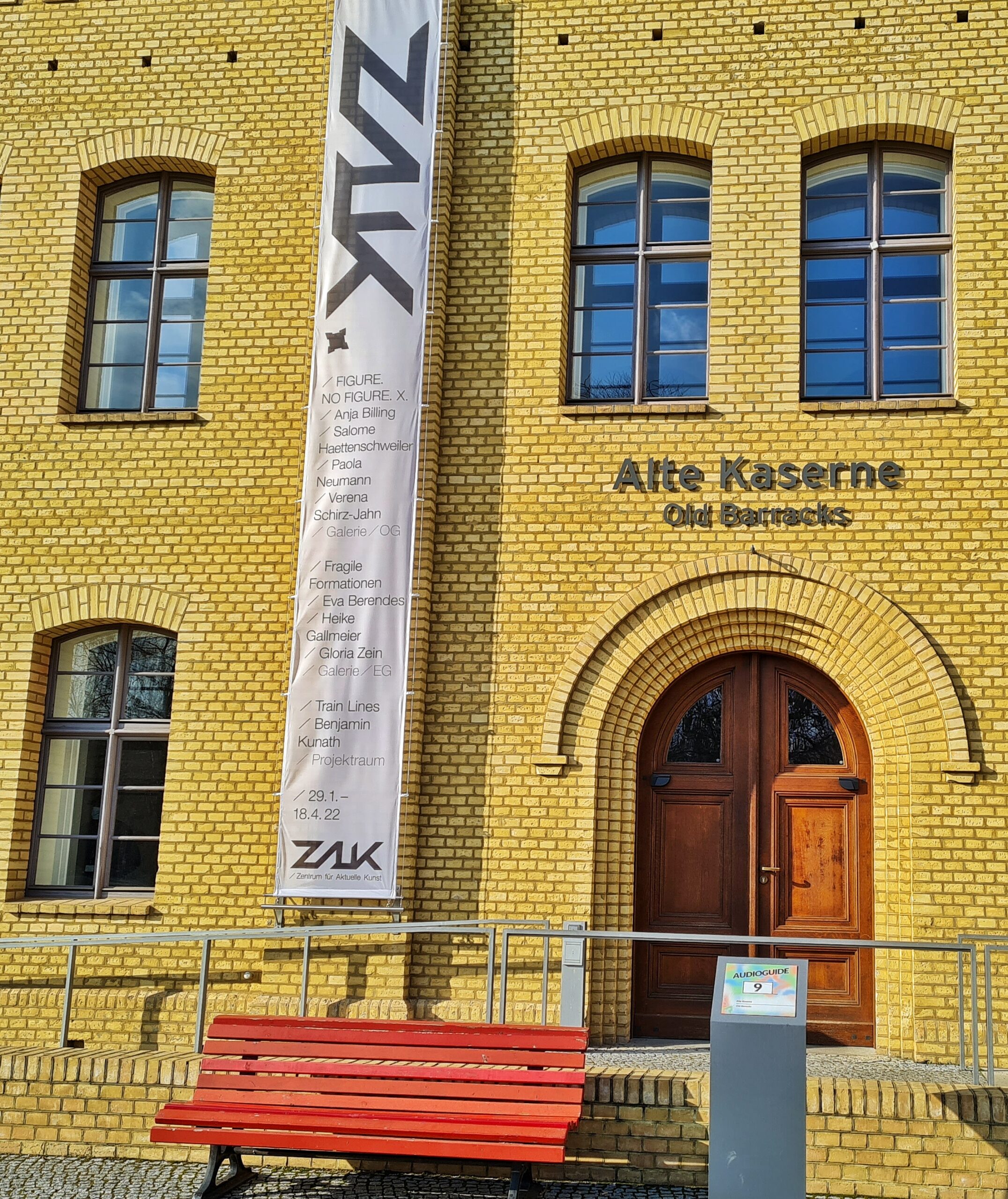 Der Eingang eines gelben Backsteingebäudes. Davor steht eine rote Bank. An dem Gebäude sind Buchstaben angebracht: "Alte Kaserne". Daneben hängt ein Banner nach unten. Darauf steht "ZAK" und die Namen von Ausstellungen mit den jeweiligen Künstlerinnen und Künstlern.