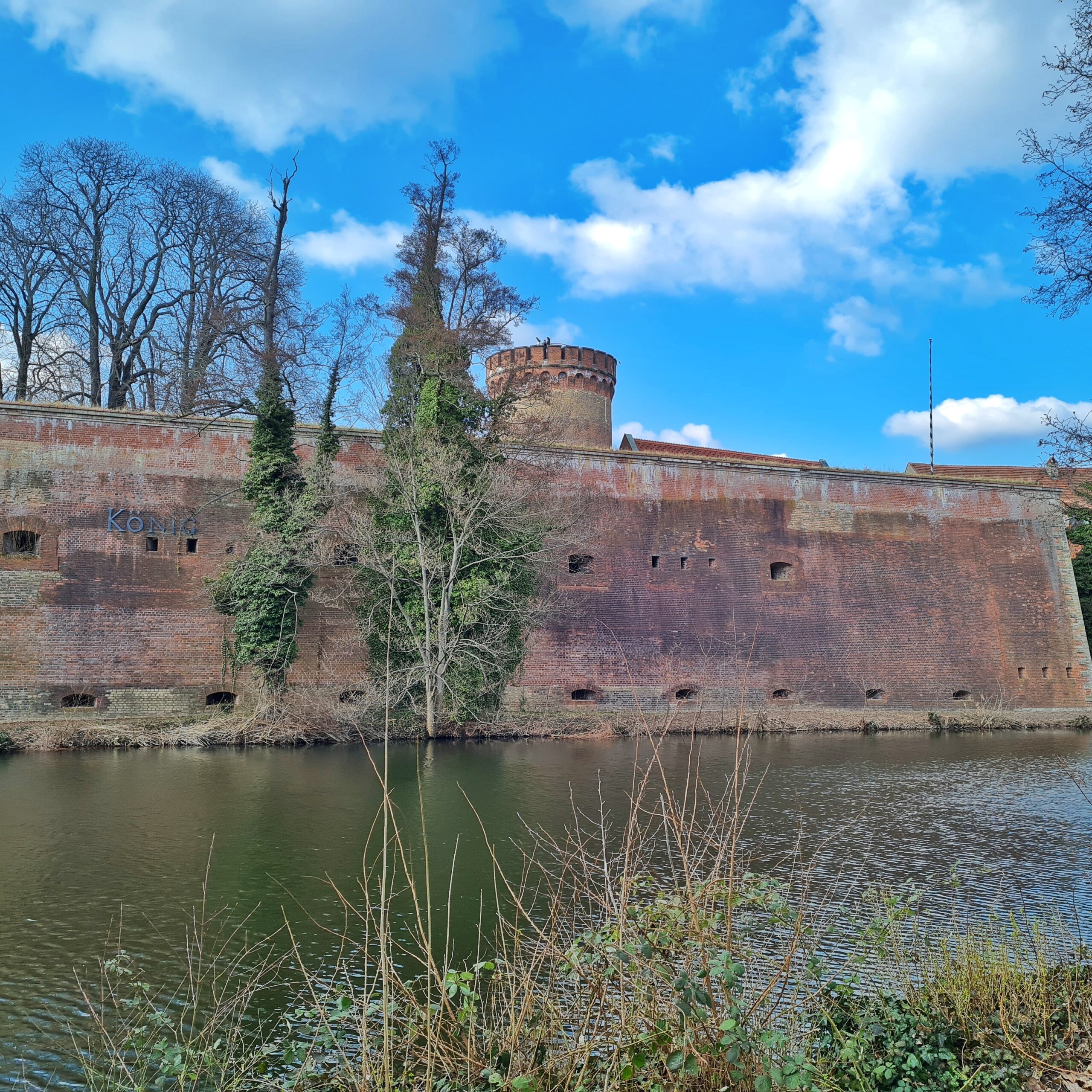 Eine hohe, massive Burgmauer, die von einem Turm mit Zinnen überragt wird. Davor eine Wasserfläche.