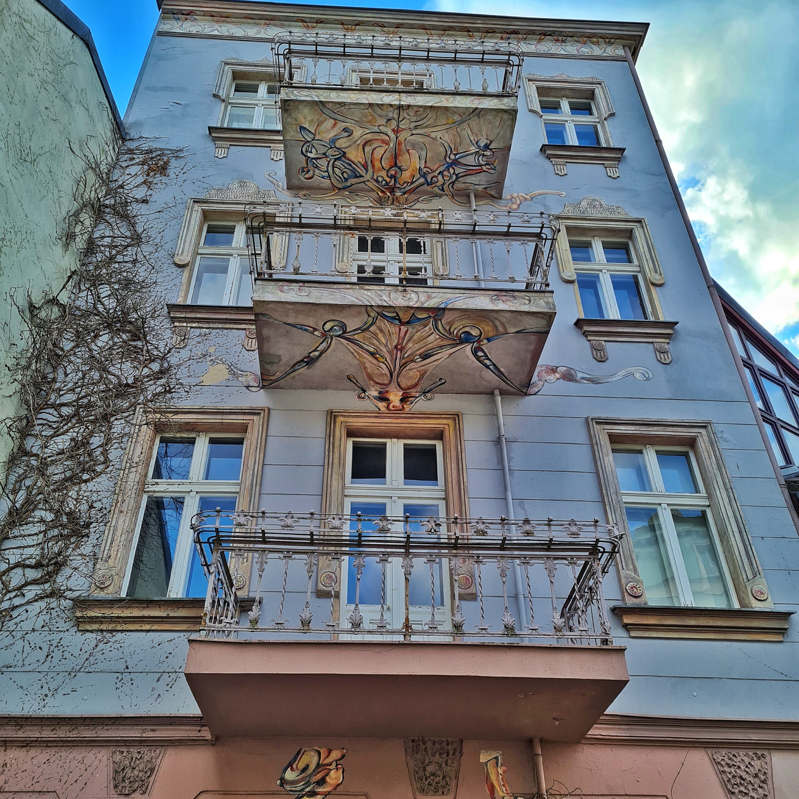 Ein schmales, dreistöckiges Haus in heller Farbe. Übereinander hat es drei Balkone. Da das Foto schräg von unten aufgenommen wurde, sieht man unter den beiden oberen Balkonen kunstvolle Bemalungen.