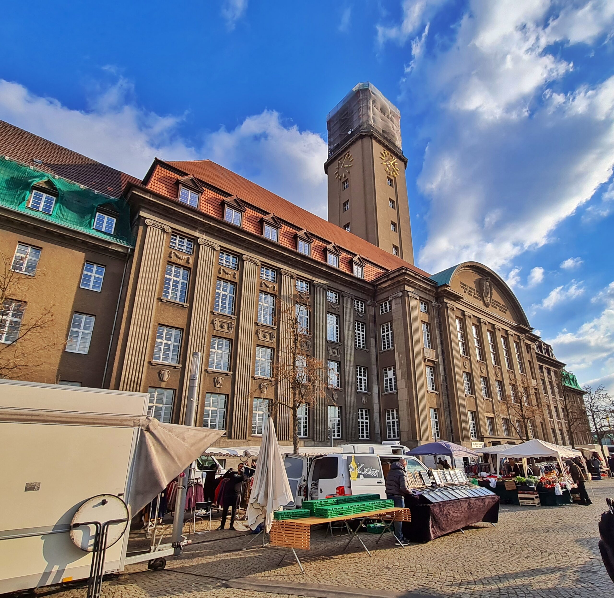 Ein sehr imposantes graues Gebäude mit vielen Fenstern neben- und übereinander. Es hat einen hohen Turm mit kleinen Fenstern und Zifferblättern. Der obere Teil des Turms ist mit einer durchsichtigen Plane bedeckt. Vor dem Gebäude stehen Marktstände.
