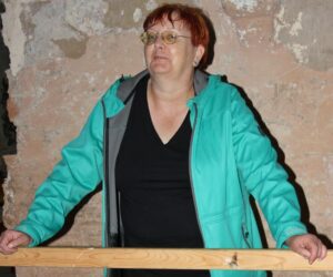 Eine mittelalte Frau mit kurzen roten Haaren, einem schwarzen Shirt und einer hellgrünen Regenjacke steht an einem Holzgeländer, auf dem sie ihre Hände abgelegt hat. Sie schaut skeptisch-verschmitzt in die Kamera.