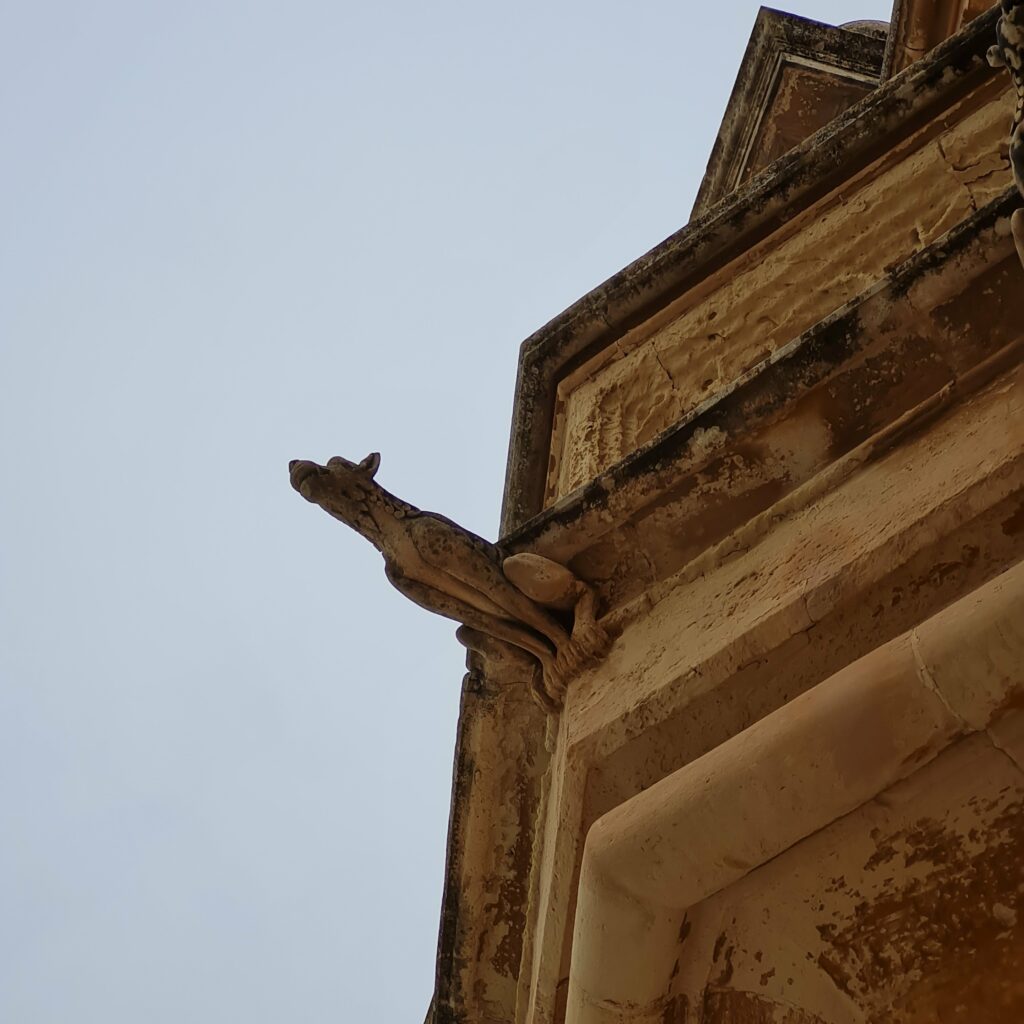 Ein bräunliches Gebäude von unten. Die Figur eines schmalen Hundes ragt vom Sims hinaus.