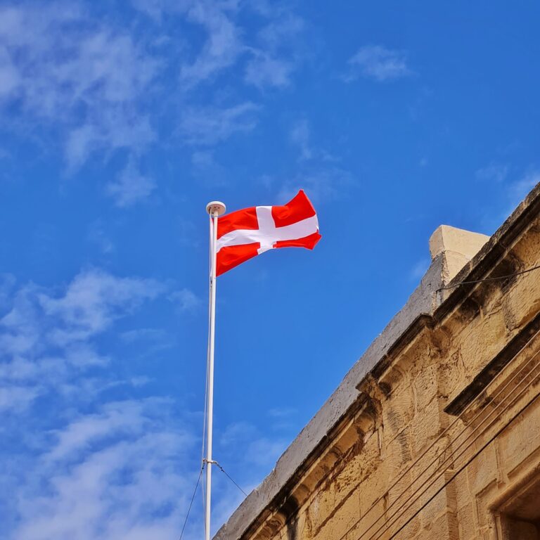 Auf einem gelblichen Gebäude, von dem nur der obere Teil erkennbar ist, weht an einer Fahnenstange vor einem blauen Himmel die maltesische Flagge (rot mit einem weißen Kreuz)