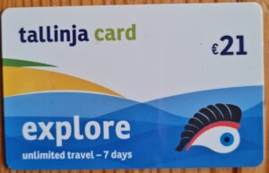 Eine Plastikkarte, auf der stilisiert blaues Meer, gelber Strrand und grüne Bäume dargestellt ist. Unten rechts ist ein großes Auge mit langen Wimpern dargestellt. Auf der Karte steht: "tallinja card", "€21", "explore", "unlimited travel - 7 days"