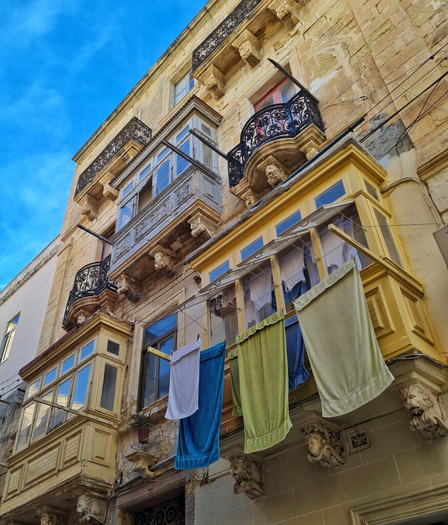 Ein Haus von unten schräg fotografiert. Es hat einige maltesische  Balkone und einige mit schmiedeeisernen Gittern. Vor einem der unteren maltesischen Balkone hängen mehrere Handtücjer in verschiedenen Farben. 