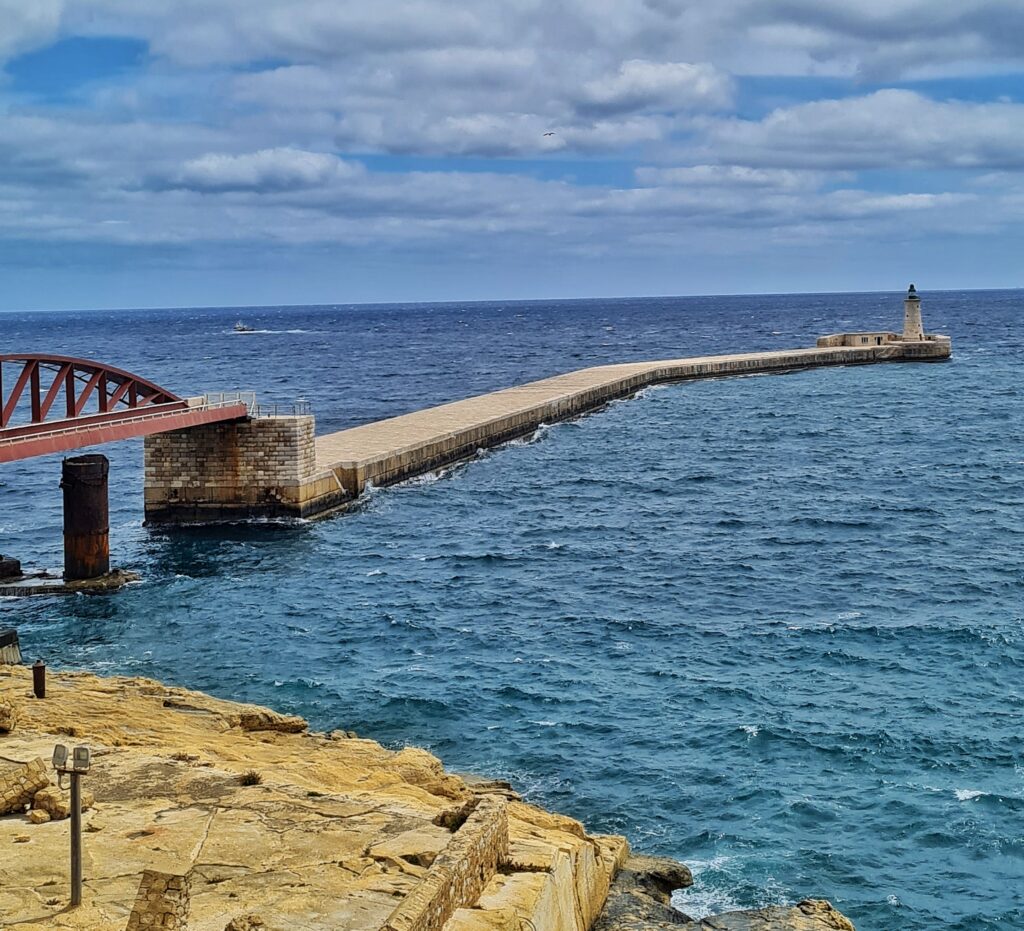 Eine lange ins Wasser ragende Mole. Am Ende ein kleiner Turm. Die Mole ist durch eine Brücke, von der man nur einen Teil sieht, mit dem Festland (von dem man nur gelbe Steine und Felsen sieht) verbunden.