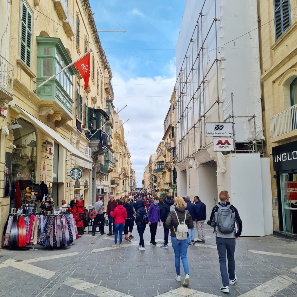 Eine schmale Straße, auf der viele Menschen laufen. Links vorne ist ein kleiner Laden, vor dem Ständern mit Schals stehen. Rechts ist ebenfalls ein Teil eines Ladens. Dahinter ist ein Gebäude mit einer Plane verdeckt. Es sind mehrere kleine Ladenschilder erkennbar, die in die Straße ragen.