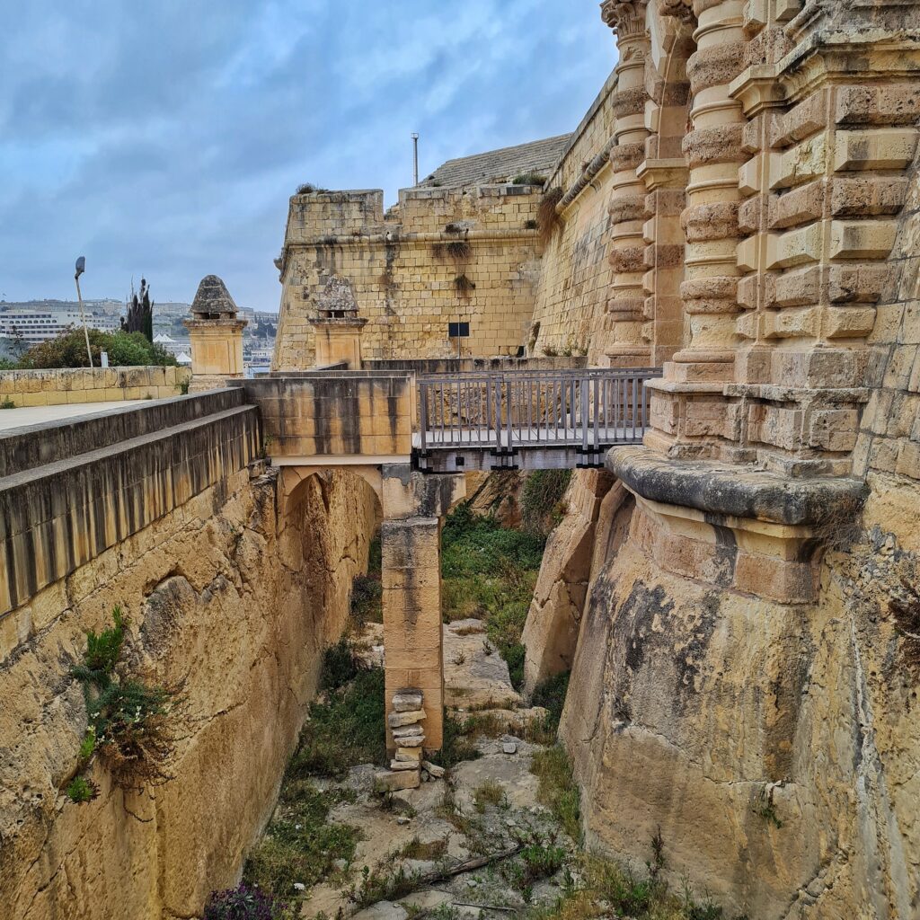 Eine Art Schlucht, die gebildet wird durch steile gelbliche Mauern. Rechts geht sie nach oben in ein gelbliches Gebäude über. Dieses setzt sich nach hinten in ein festungsartiges Gebäude fort. Auf der linken Mauer ist oben eine gerade Fläche. Über die Schlucht geht eine kleine Brücke, die unten von einem großen Pfeiler getragen wird.