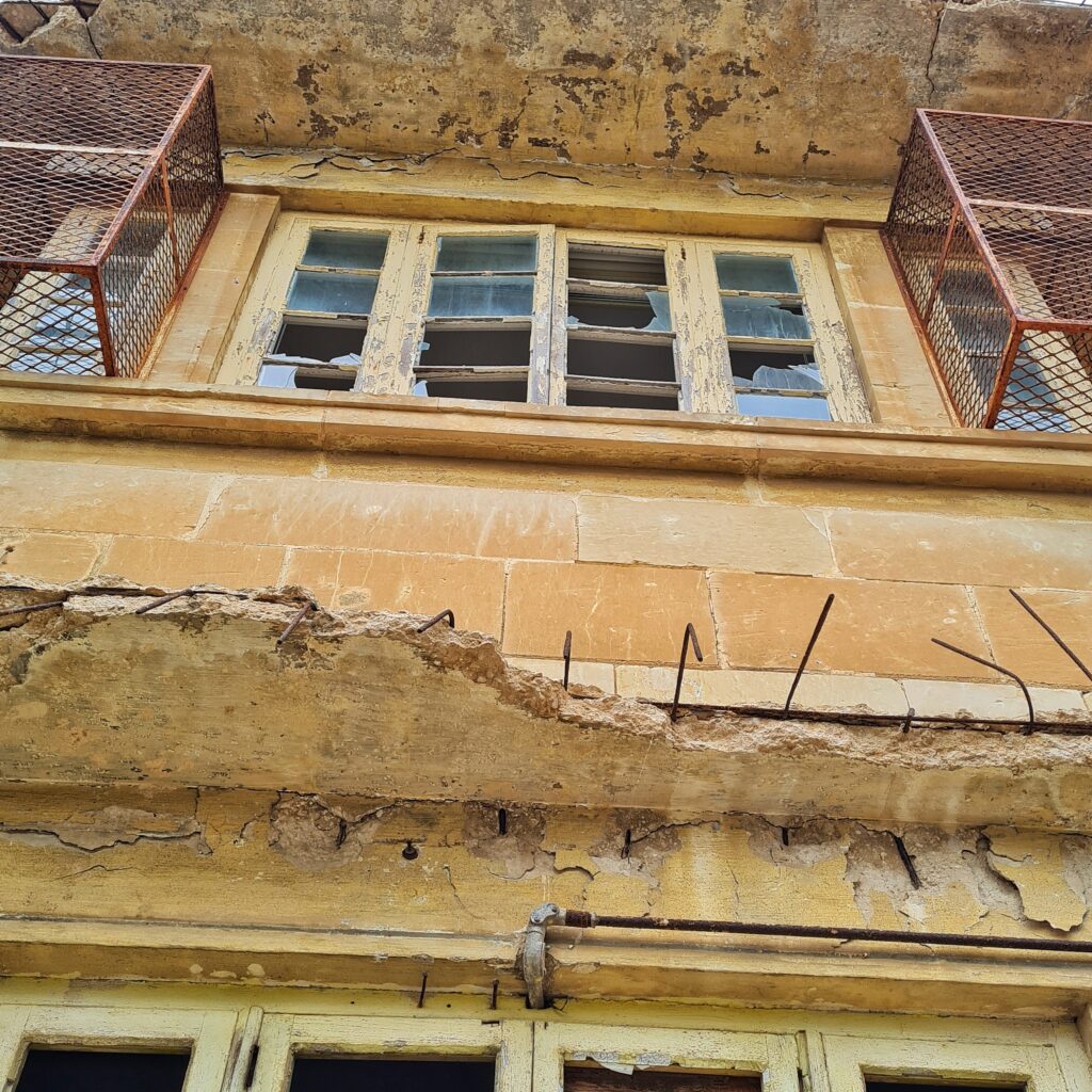 Ein Teil eines gelblichen Hauses schräg von unten fotografiert. Unten sieht man den oberen Teil von leeren Fensteröffnungen. Darüber ein Überstand, der bröckelig ist. Mehrere kleine Metallstangen ragen hervor. Ganz oben rechts und links zwei schmale Fenster, vor denen rostige Gitterkästen angebracht sind. In der Mitte ein breites Sprossenfenster bei dem sie meisten Scheiben ganz fehlen oder nur Glassplitter vorhanden sind.