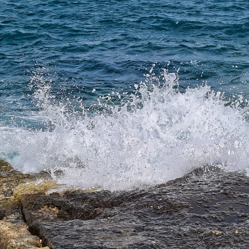 Im oberen Bildteil dunkelblaues welliges Wasser. Unten ein flacher, nasser Felsen, auf den im mittleren Bildteil Wasser mit hochspritzender weißer Gischt auftrifft.