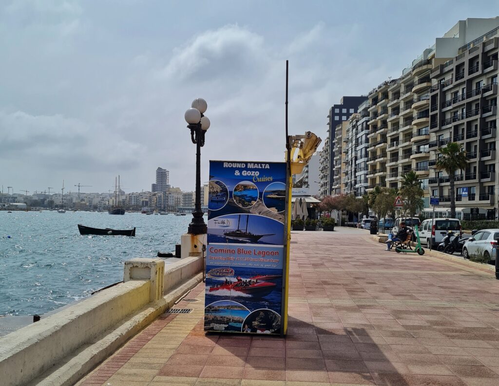 Eine breite Promenade. Links Wasser, das in der Sonne glitzert. Ganz im Hintergrund sind Schiffe und Gebäude zu sehen. iRechts eine hohe Häuserreihe. Die Gebäude sind modern und haben viele Balkone. Zwischen den Häusern und der Promenade eine Straße mit Bäumen und parkenden Autos. Auf der Promenade steht ein großes Plakat, das mit Fotos für Schifffahrten wirbt.
