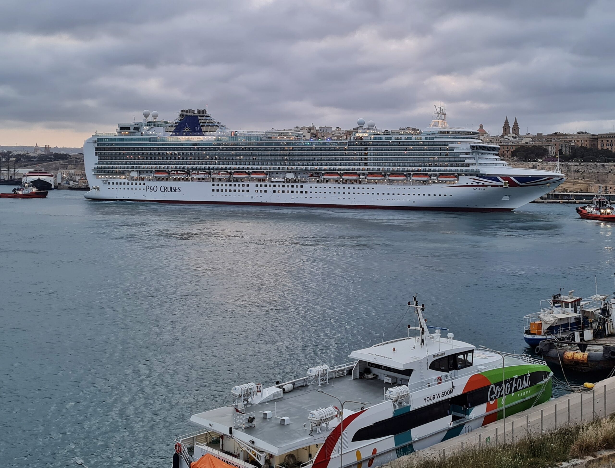 Ein weißes Kreuzfahrtschiff mit der Aufschrift "P & O Cruises" liegt am Kai vor Valletta. Vor dem Bug ein roter Schlepper. Auf der Seite des Hafens, von wo das Foto gemacht wurde, liegen eine Fähre mit den Aufschriften "Gozo Fast" und "Your Wisdom". Davor liegt ein anderes schwarzes Boot.