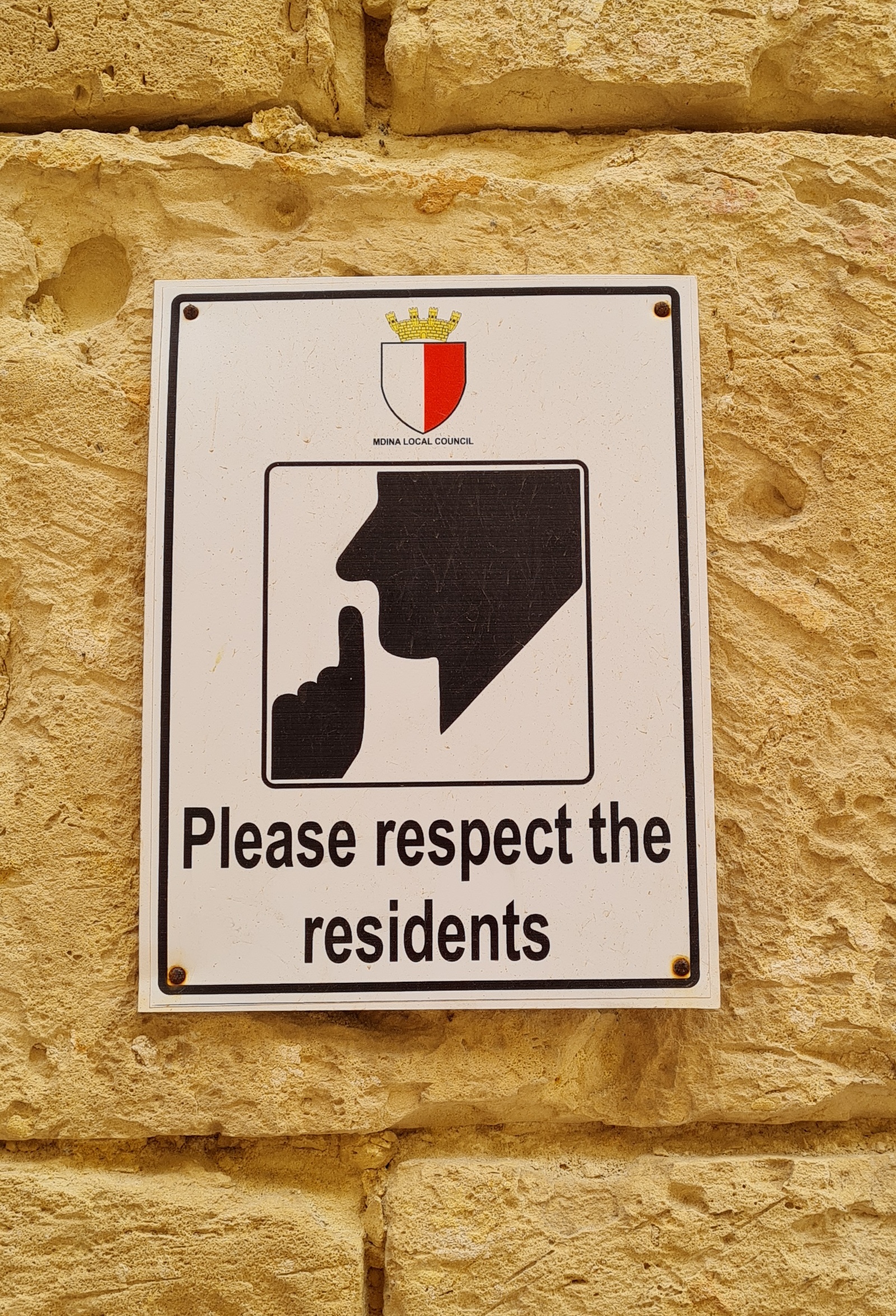 Ein weißes Schild an einer Wand. Darauf stilisiert ein Kopf von der Seite mit einem erhobenen Zeigefinger vor dem Mund. Darunter steht "Pleace respect the residents". Oben auf dem Schild ist ein kleines Wappen (ein Schild, links weiß, rechts rot, und darüber eine Krone). Darunter steht in sehr kleiner Schrift "Mdina Local council"
