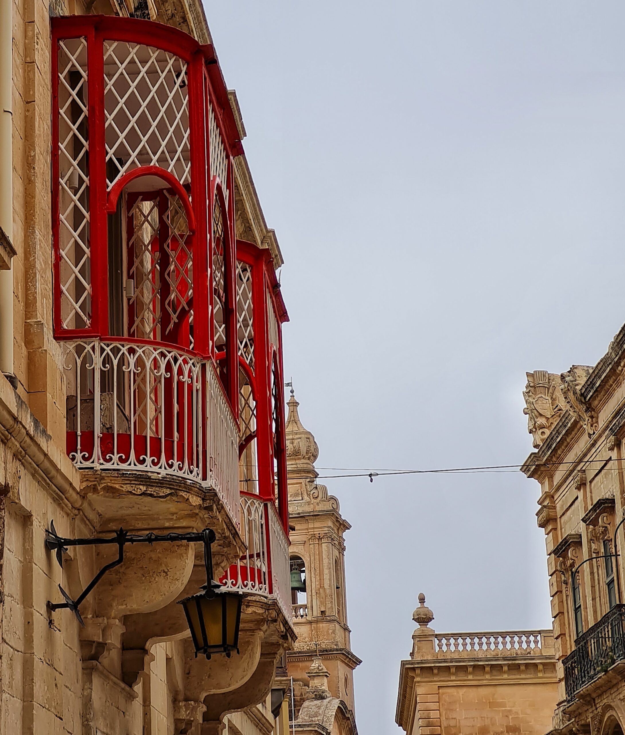 Links in offener Balkon mit weißen Gittern und roten Holzstreben. Im Hintergrund ein Kirchturm. Rechts ein Teil eines hohen, hellen Gebäudes.