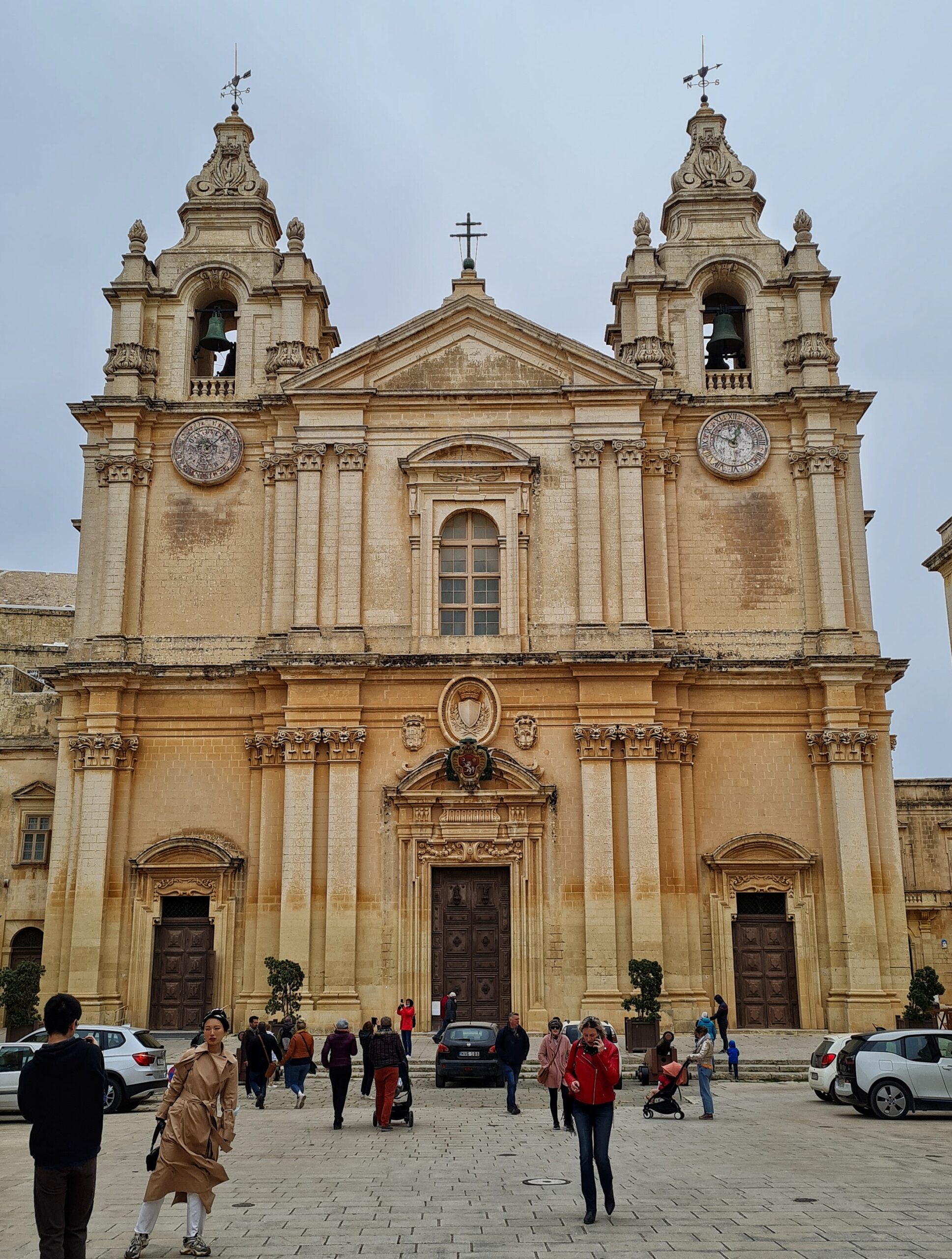 Eine große, helle Kirche mit zwei Türmchen im Hintergrund eines Platzes. Auf dem Platz stehen wenige Autos, und es laufen Menschen darüber. Links unten posiert eine Frau im Mantel für ein Foto.