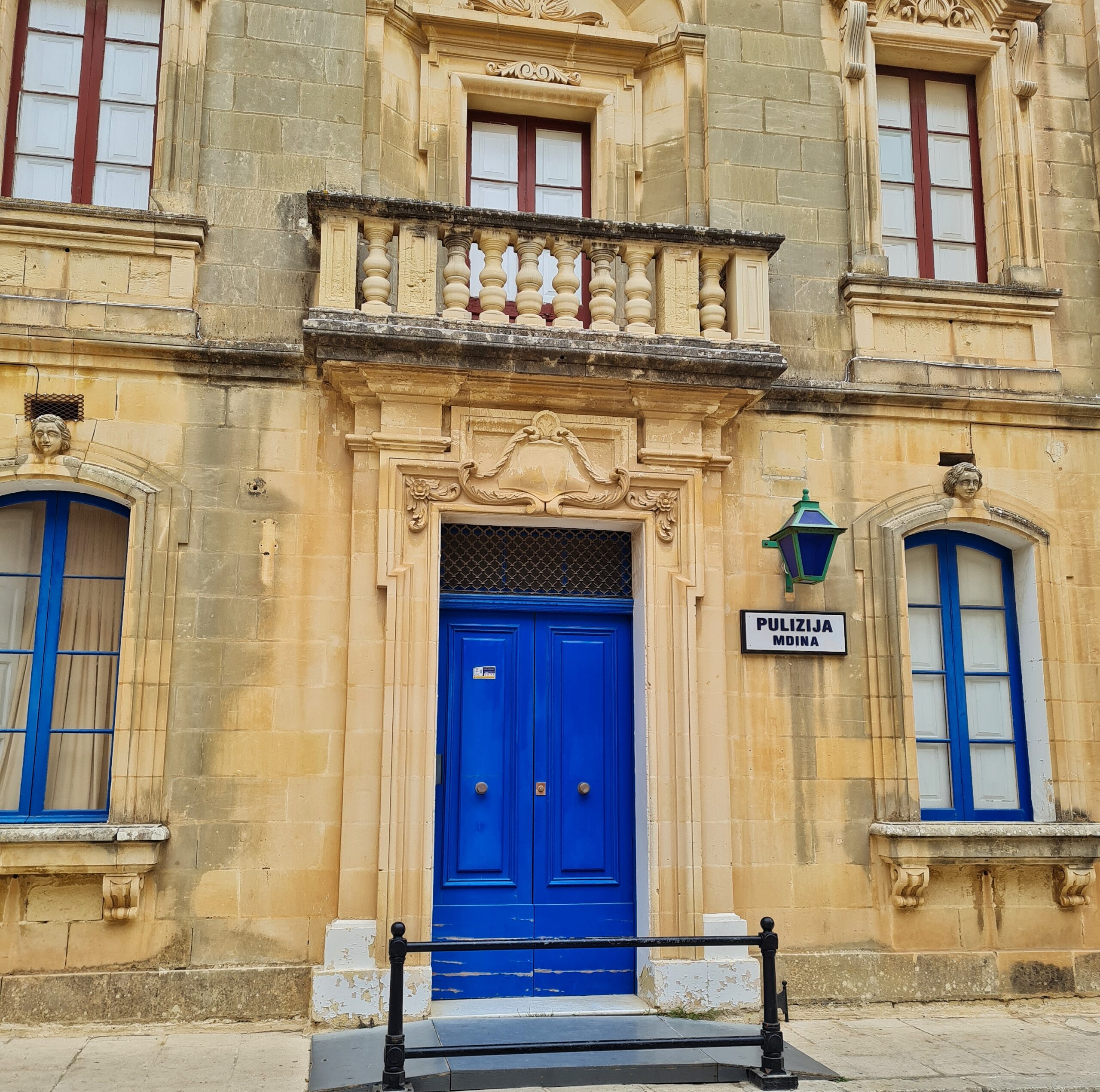 Ein gelbliches Gebäude mit einer blauen Tür, daneben ein Schild: "Pulizija Mdina". Darüber ein Balkon. Die Fenster unten haben blaue Sprossen, die Fenster oben rote.