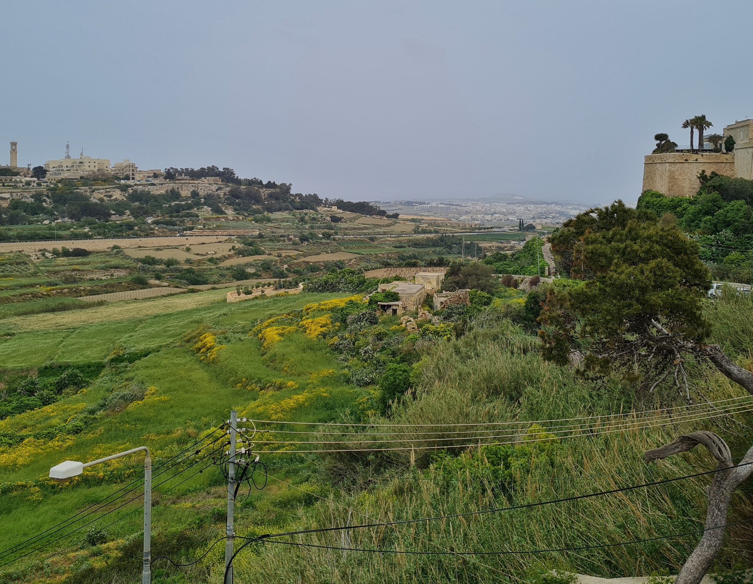 Ein weiter Blick in eine grüne Landschaft. Ganz im Hintergrund ist unscharf eine Stadt mit vielen Gebäuden zu erkennen. Links liegt erhöht eine Ortschaft mit größeren Gebäuden und hohen Türmen. Rechts im Bild sind Teile einer Festung und hohe Bäume erkennbar.