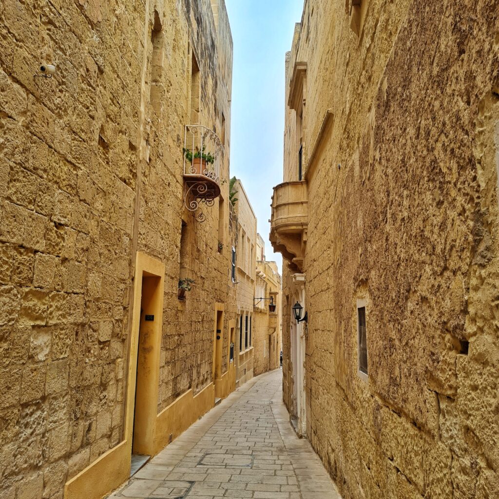 Eine ganz schmale Gasse, begrenzt durch hohe, gelbliche Gebäudewände, an denen teilweise Balkone, Fenster und Türeingänge erkennbar sind.
