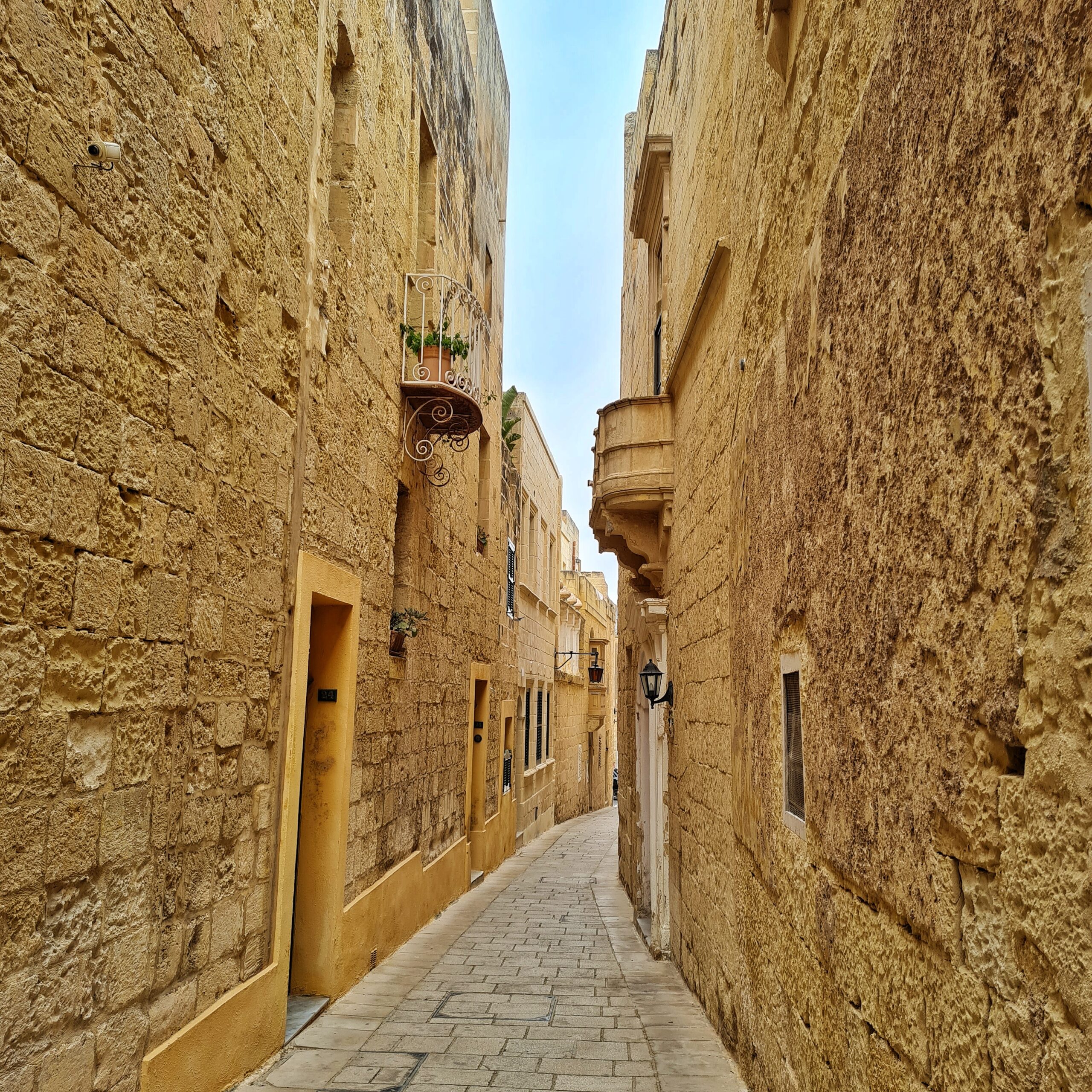 Eine ganz schmale Gasse, begrenzt durch hohe, gelbliche Gebäudewände, an denen teilweise Balkone, Fenster und Türeingänge erkennbar sind.