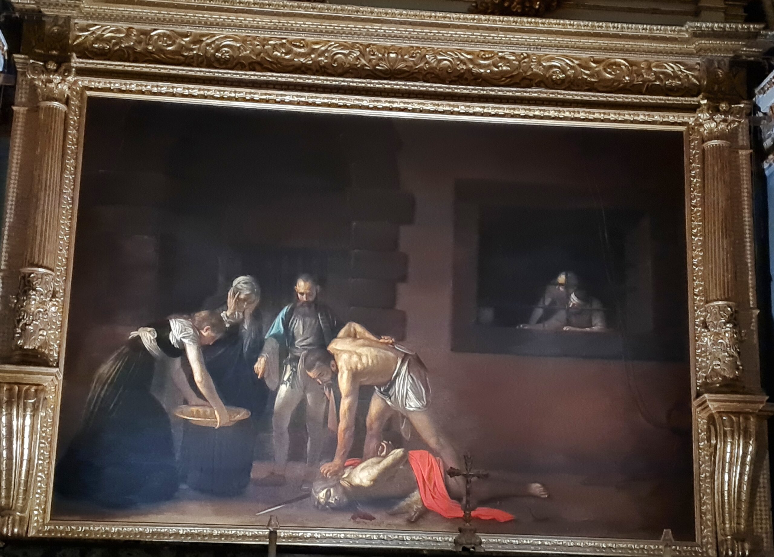 Ein sehr großes Gemälde. Im linken, dominierenden Bildteil liegt ein Mann, der nur ein rotes Tuch um die Lenden trägt, auf dem Boden. Vor ihm ist Blut zu sehen und hinter ihm die Schneide eines Schwertes. Ein Mann beugt sich zu ihm herunter und packt ihn am Kopf. Von links kommt eine junge Frau, die gebeugt eine große Schale in beiden Hämden trägt. Dazwischen eine alte Frau die entsetzt die Hände vors Gesicht schlägt und ein alter Mann, der auf die Schale zeigt. Rechts ist ein vergittertes Fenster zu sehen, durch das zwei Personen die Szene beobachten.