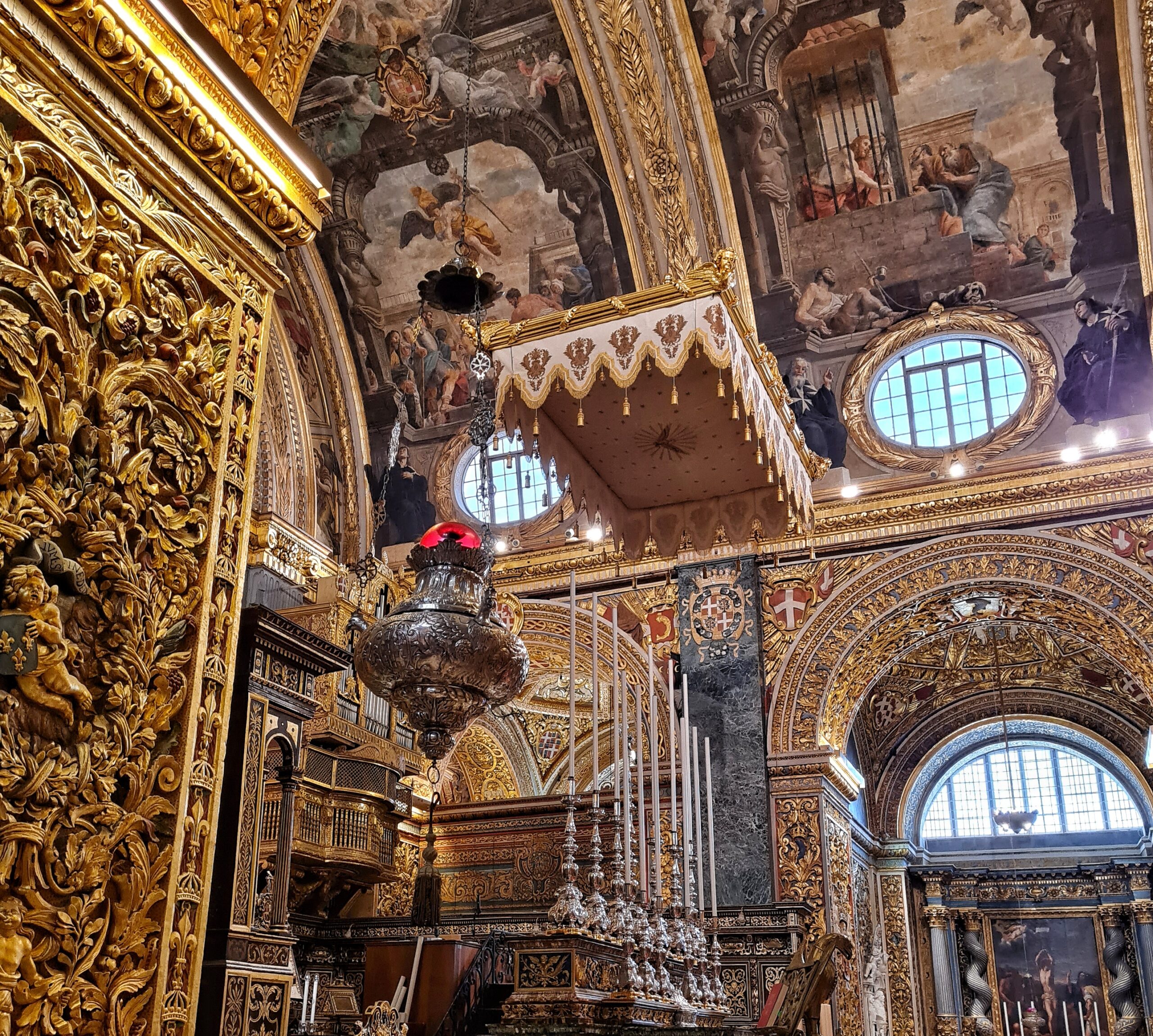 Blick auf einen Altar, über dem ein weiß-goldener Baldachin hängt. Davor stehen sehr hohe weiße Kerzen in silbernen Leuchtern, und von der Decke hängt ein silbernes großes Gefäß. Die Wände und Decken sind in Gold und mit Gemälden reich verziert.