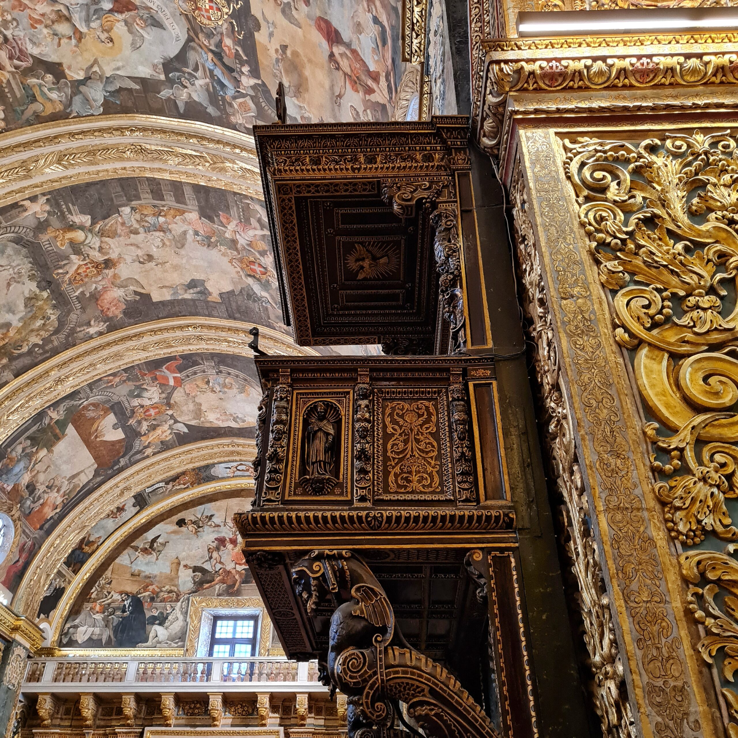 Eine Kanzel aus dunklem Holz, reich verziert; seitlich von unten fotografiert. Darüber ist unscharf ein buntes Deckengemälde zu sehen und rechts ein Teil einer Wand mit goldenen Ornamenten.