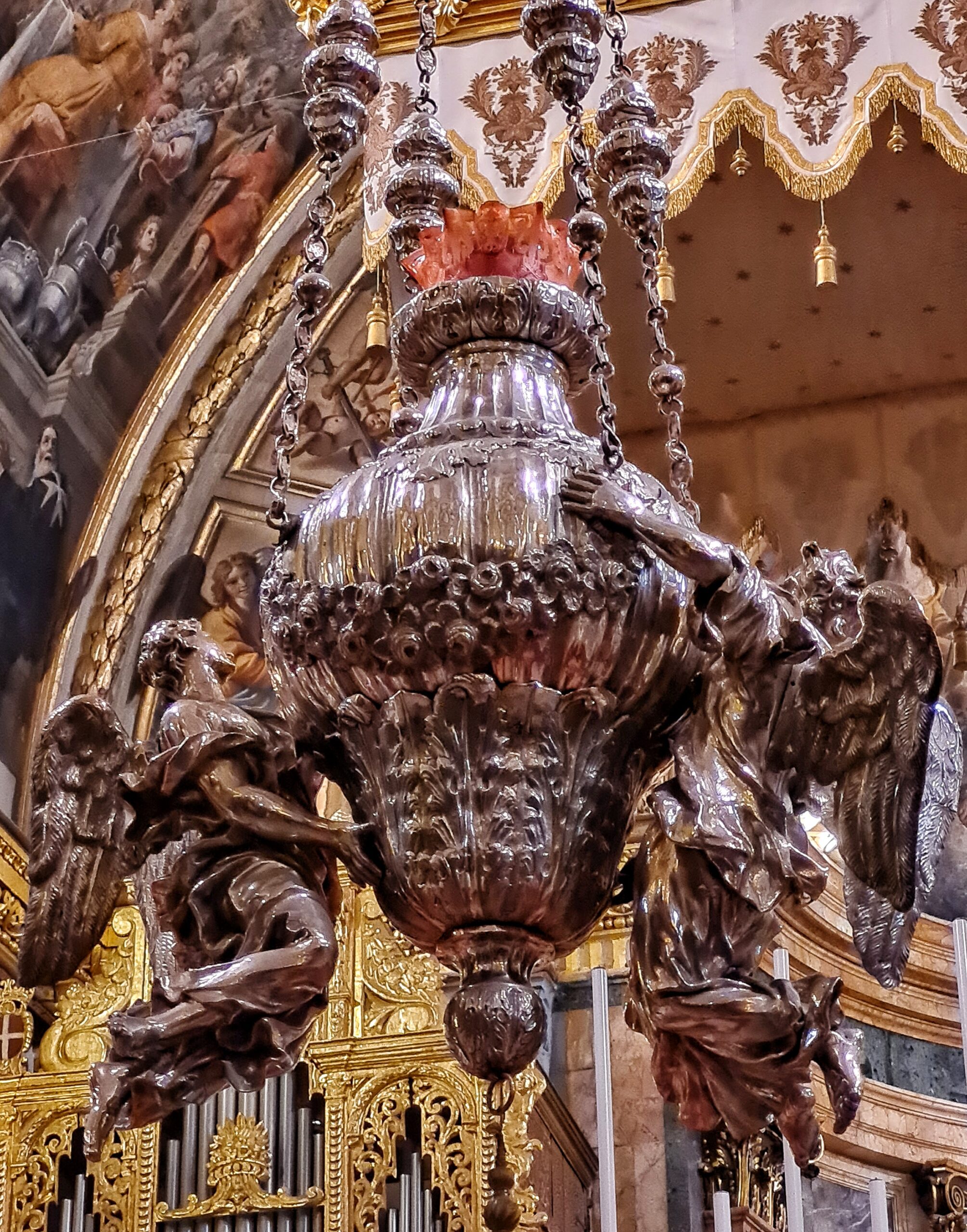 Ein großesdunkel-silbernes Gefäß, das reich verziert ist. Oben sind verzierte Metallbänder zu sehen, an den das Gefäß offenbar hängt. Links und rechts an dem Gefäß hängen zwei kunstvoll gearbeitete fliegende
Engel. Sie sind so dargestellt, als ob sie das Gefäß tragen würden.