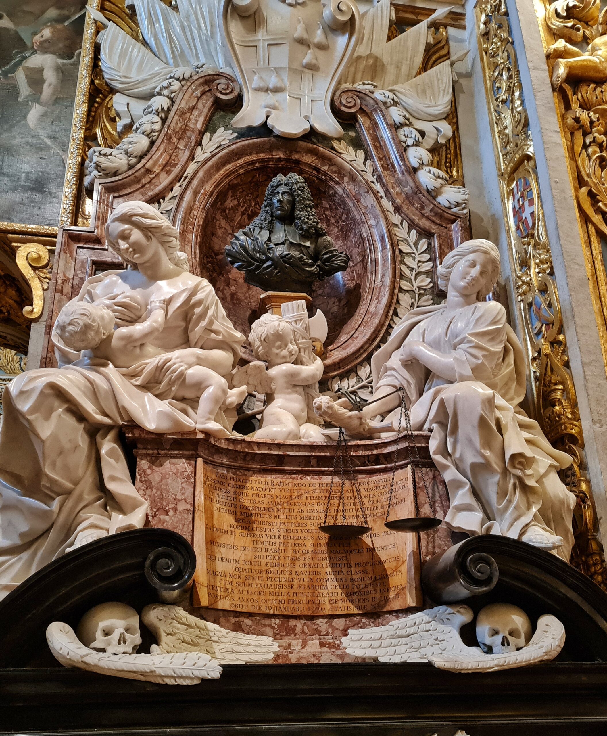 In einer runden Nische die dunkle Büste eines Mannes mit langem, gelockten Haar. Darum weiße Verzierungen. Davor drei weiße Statuen: links eine sitzende Frau, die ein Kind auf dem hält, rechts eine sitzende Frau, über deren Arm eine Waage hängt. In der Mitte ein kleiner sitzender Engel. Darunter links und rechts zwei Totenkopfdarstellungen.