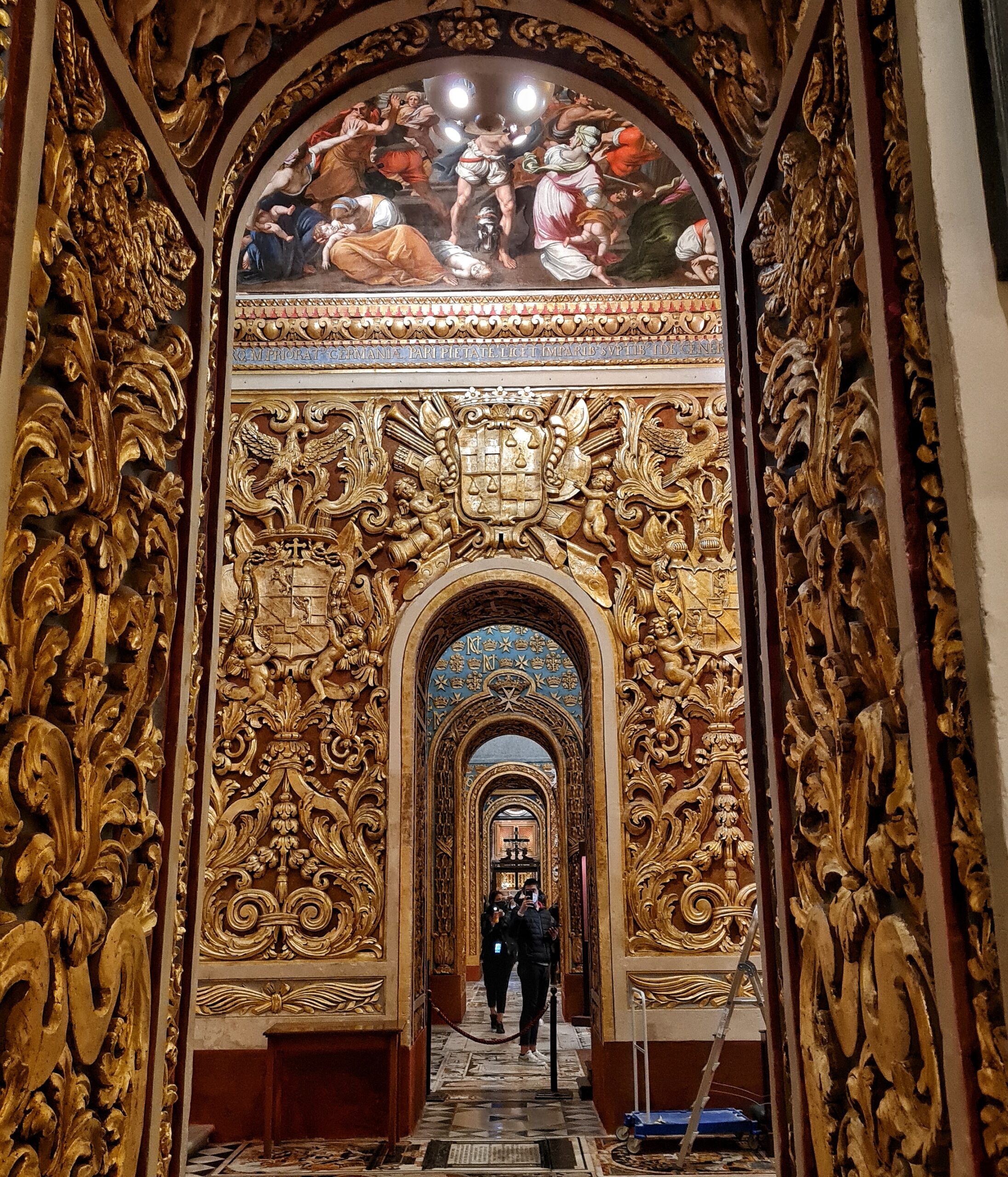 Blick durch eine Reiihe von länglichen Öffnungen, die oben halbrund abschließen. Im Hintergrund sind Menschen zu sehen. Die vordere öffnung ist mit goldenen Ornamenten reich verziert. Ebenso die Wand dahinter. Darüber ist ein halbrundes Gemälde.