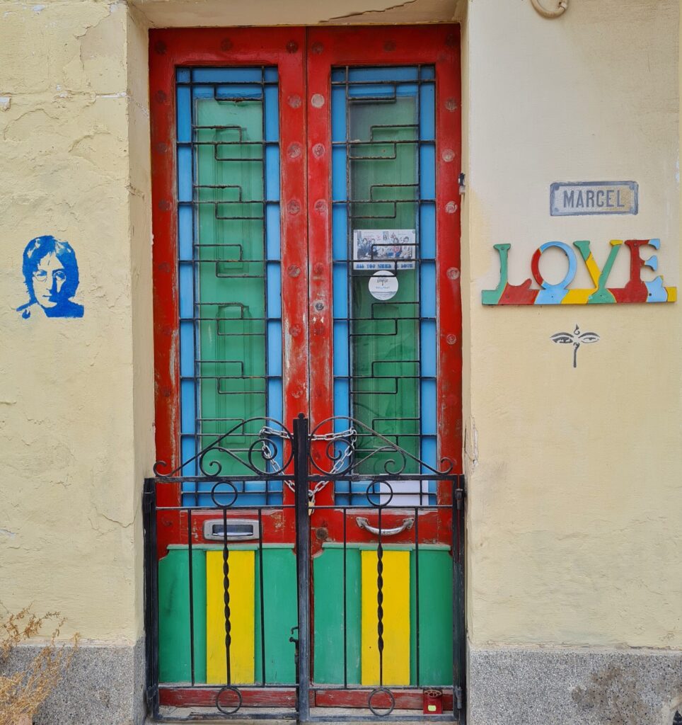 Eine vergitterte Haustür. Sie ist in rot, türkis, hellblau und gelb gestrichen. Im oberen Bereich hängt eine Postkarte, auf der mehrere Menschen zu sehen sind und der Text "All you need is love". Links von der Tür eine Graffitizeichnung, die den Kopf von ohn Lennon zeigt. Rechts von der Tür ein Schild mit der Aufschrift "Marcel". Darunter bunt bemalt die Buchstaben "LOVE". Darunter eine Zeichnung, die zwei Augen mit angedeuterter Nase und einem Symbol darüber zeigt. 
