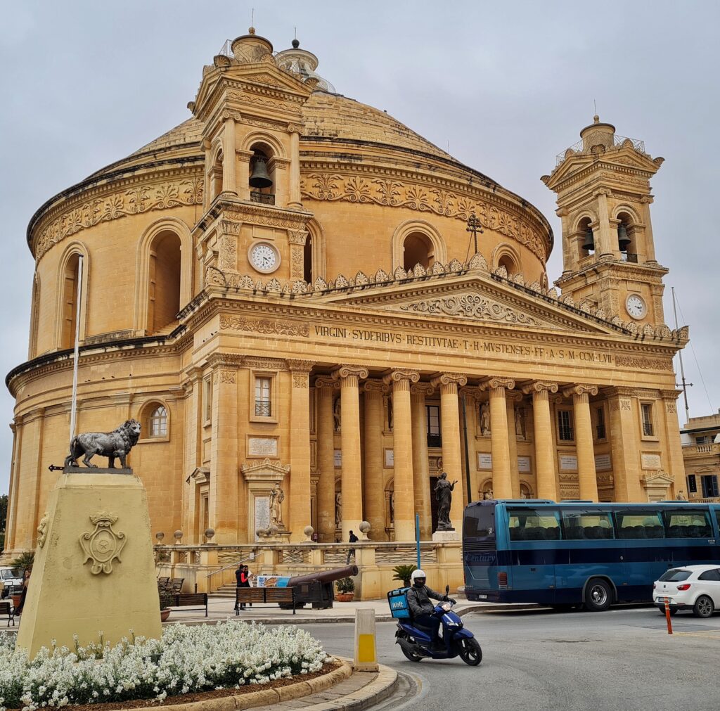 Eine Kirche aus gelblichem Stein. Im Eingangsbereich mehrere Säulen und links und rechts zwei Türme, oben Glocken, darunter Uhren. Der hintere Teil der Kirche ist rund und hat ein kuppelförmiges Dach. Vor der Kirche ein Denkmal mit einem Löwen darauf und fahrende Fahrzeuge (ein Bus, ein Pkw, ein Motorrad)