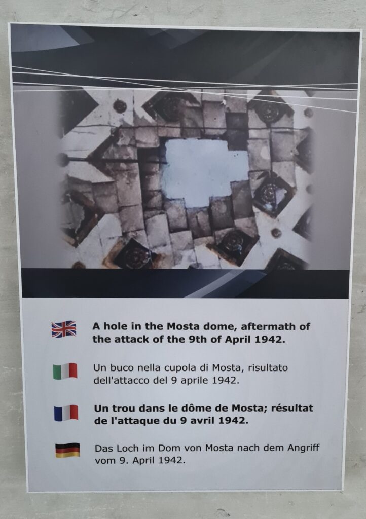 Ein Foto von einem Loch in einer Steindecke. Darunter steht Text in Englisch, Italienisch, Französisch und Deutsch. Der deutsche Text lautet "Das Loch im Dom von Mosta nach dem Angriff vom 9. April 1942".