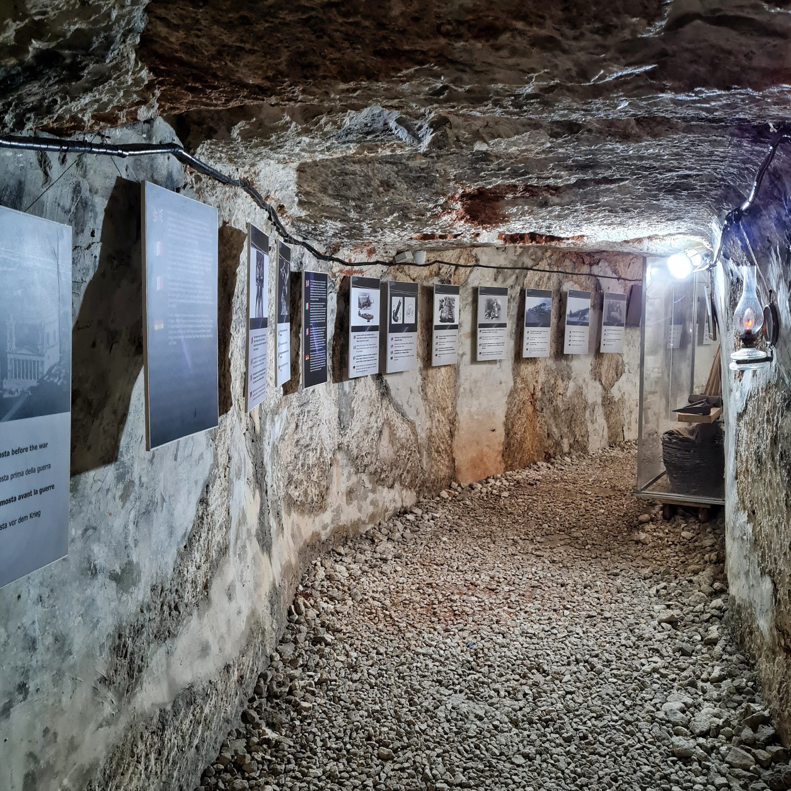 Ein beleuchteter Bunkergang mit steinigem Boden und einer niedrigen felsigen Decke. An den Wänden hängen Fotos mit Text.