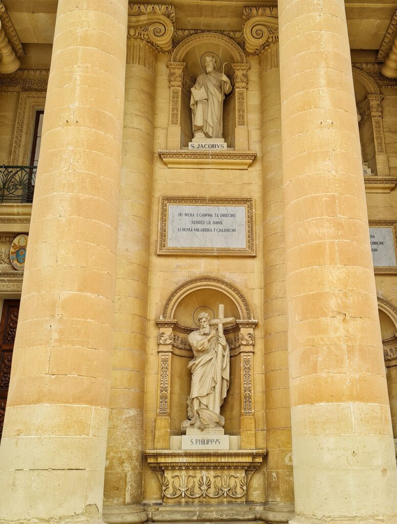 Hohe Säulen aus gelblichem Stein bilden eine Nische. In dieser sind zwei weitere Nischen, untereinander. In diser steht jeweils eine Figur in langen Gewändern und mit Heiligenschein. Die untere zeigt einen Mann, der ein  Kreuz trägt, die obere einen Mann mit Bischofsstab. In der anderen Hand hält er ein Blatt Papier.