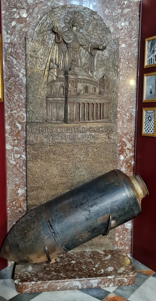 Eine Bombe vor einer hohen Metallplatte. Auf dieser ist oben das Bild einer Kirche mit kuppelförmigen Dach und einer großen Figir eingeritzt. Die Figur trägt ein langes Gewand und hat um den Kopf einen Strahlenkranz. Sie hält die Hände ausgebreitet über die Kirche. Darunter steht "IL-Miraklu Tal-Bomba" " 9 Ta April 1942". 