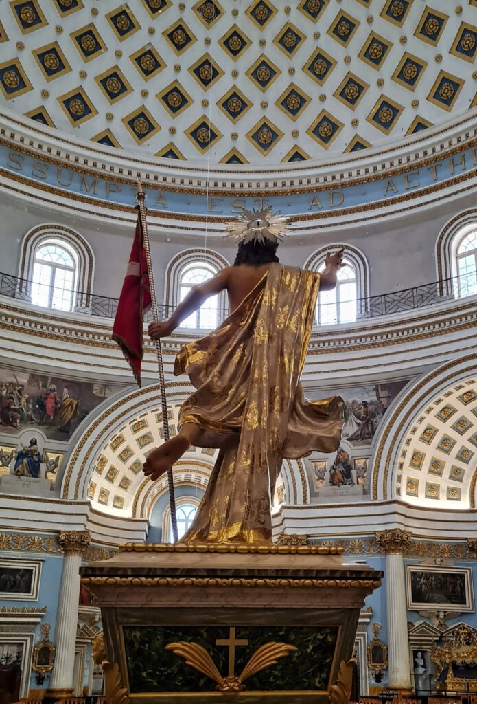 Die Christusstatue von hinten fotografiert. Die linke Fußsohle ist in Richtung Kamera gestreckt. Im Hintergrund weiße Wände und Bögen mit goldenen Mustern und Gemälden. Oben eine Reihe von Fenstern und darüber ein Teil der viereckigen Muster der Kuppel.