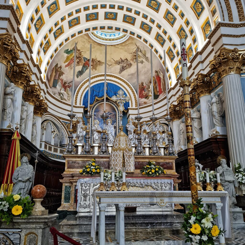 Ein Altar in weiß,  kt gelben und weißen Blumengestecken geschmückt. Er ist umrahmt von weißen Pfeilern, oben mit Gold verziert, und weißen Statuen. Oben ist ein Bogen mit blau-goldene Mustern und ein Deckengemälde, das mehrere Menschen und Engel in farbigen Gewändern zeigt.