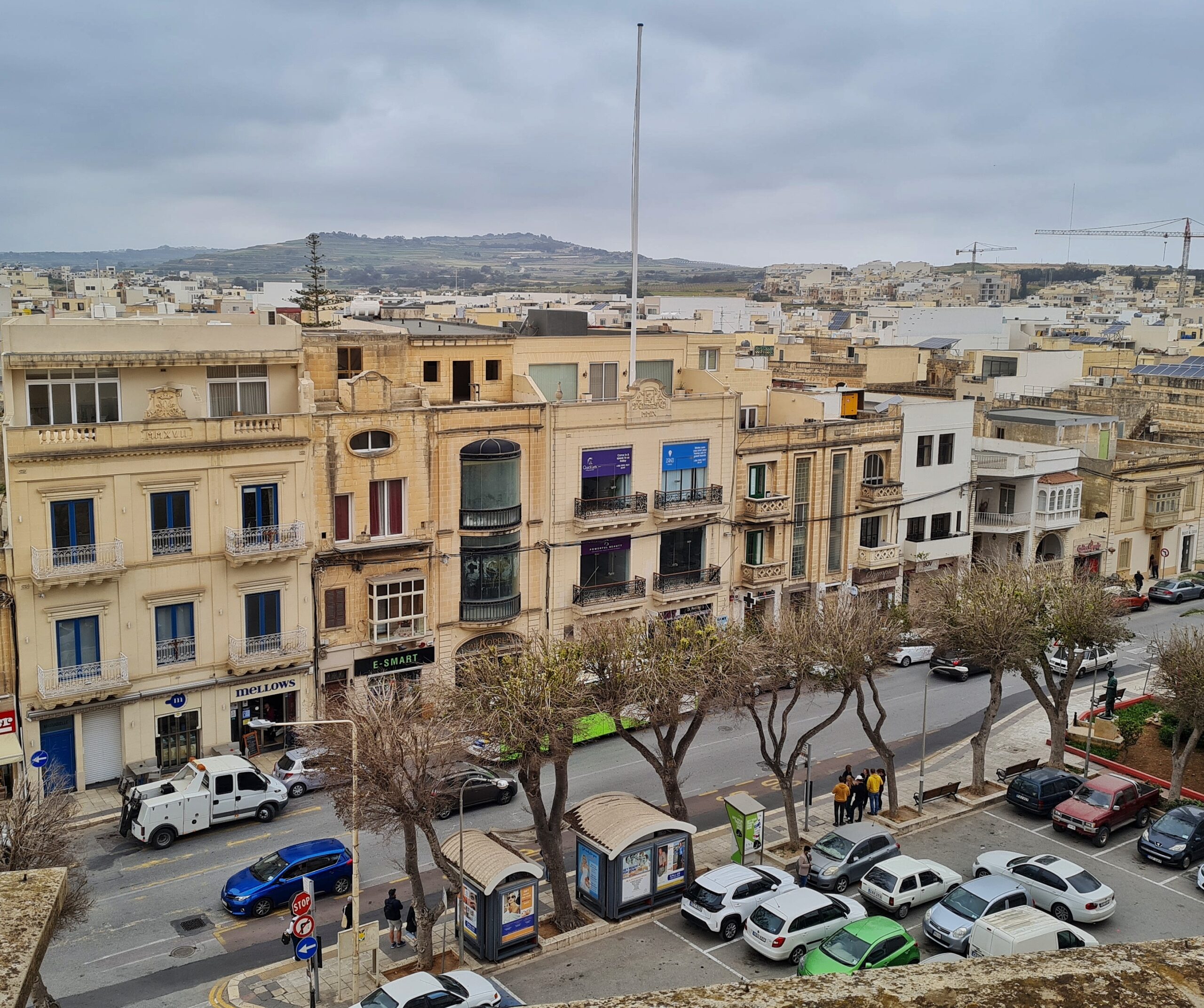 Blick auf eine Straße von schräg oben fotografiert. Auf der Straße fahren und parken einige Autos. Im unteren Bildbereich ist eine Bushaltestelle, an der Menschen warten, und ein Parkplatz, auf dem Autos stehen. Auf der anderen Straßenseite eine dreigeschossige, durchgehende Häuserreihe. Dahinter weitere Bebauung und ganz im Hintergrund ein flacher Hügel.