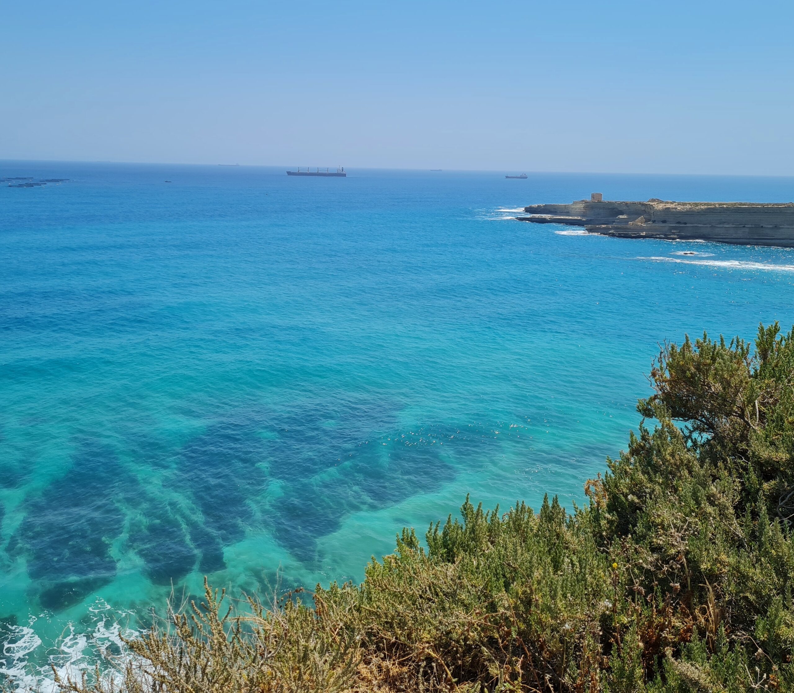 Das Meer von einem höher gelegenen, mit niedrigen Büschen bewachsenen Standort aus fotografiert. Das Meer ist blau-grün und ganz klar, so dass man bis auf den Grund mit dunklen Flecken sehen kann. Rechts hinten ist eine schmale Landzunge zu erkennen und im Hintergrund ganz in der Ferne zwei Frachtschiffe.