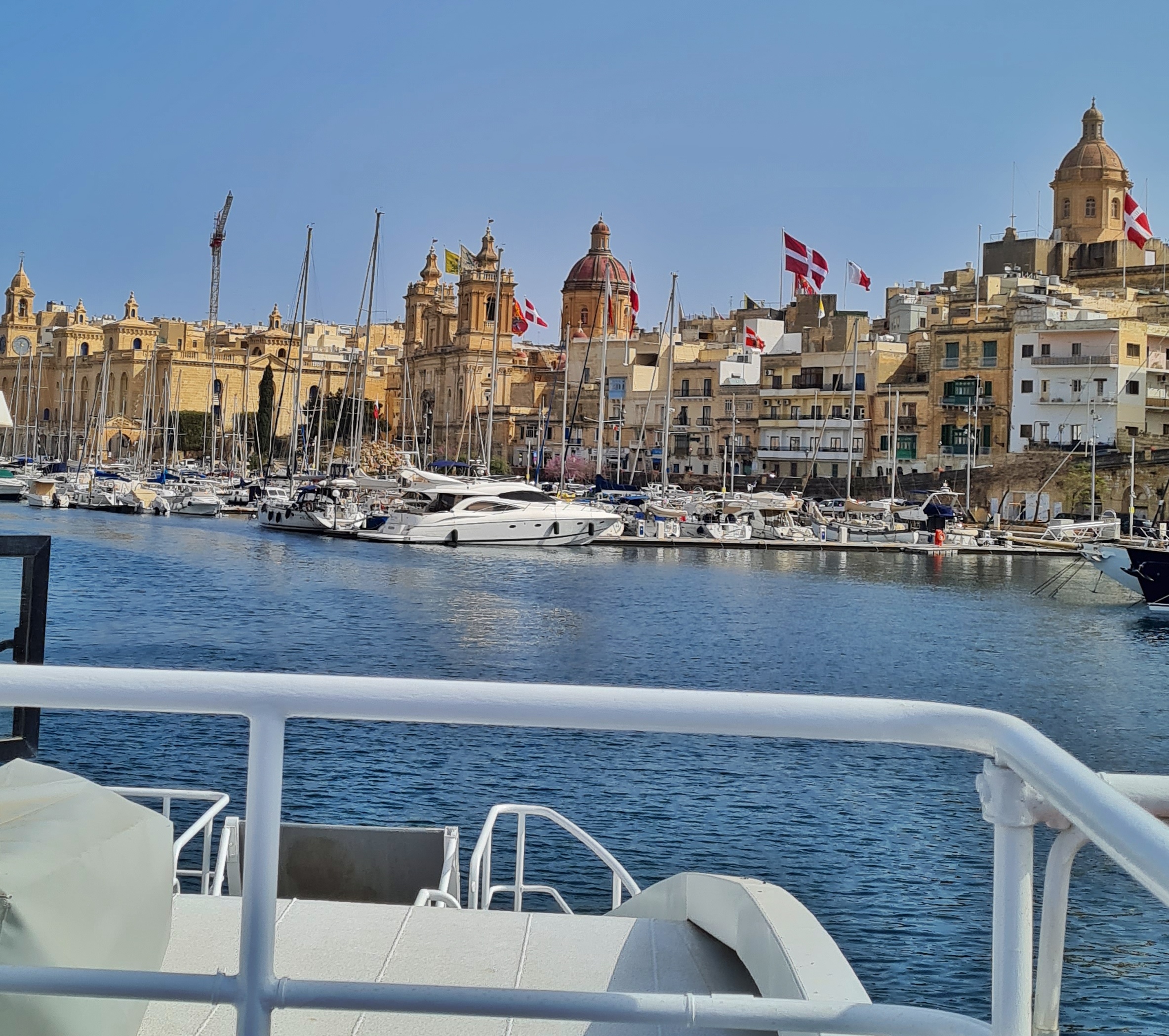 Im Vordergrund eine Reling und ein Teil eines kleineren, weißen Schiffes. Dahinter Wasser, und auf der anderen Seite der Wasserfläche viele Segelboote und Yachten an Anlegestegen, eine Festung, eine Kirche, zwei Türme und hohe Häuser. Alle Gebäude sind gelb, beige, leicht rötlich oder ocker.