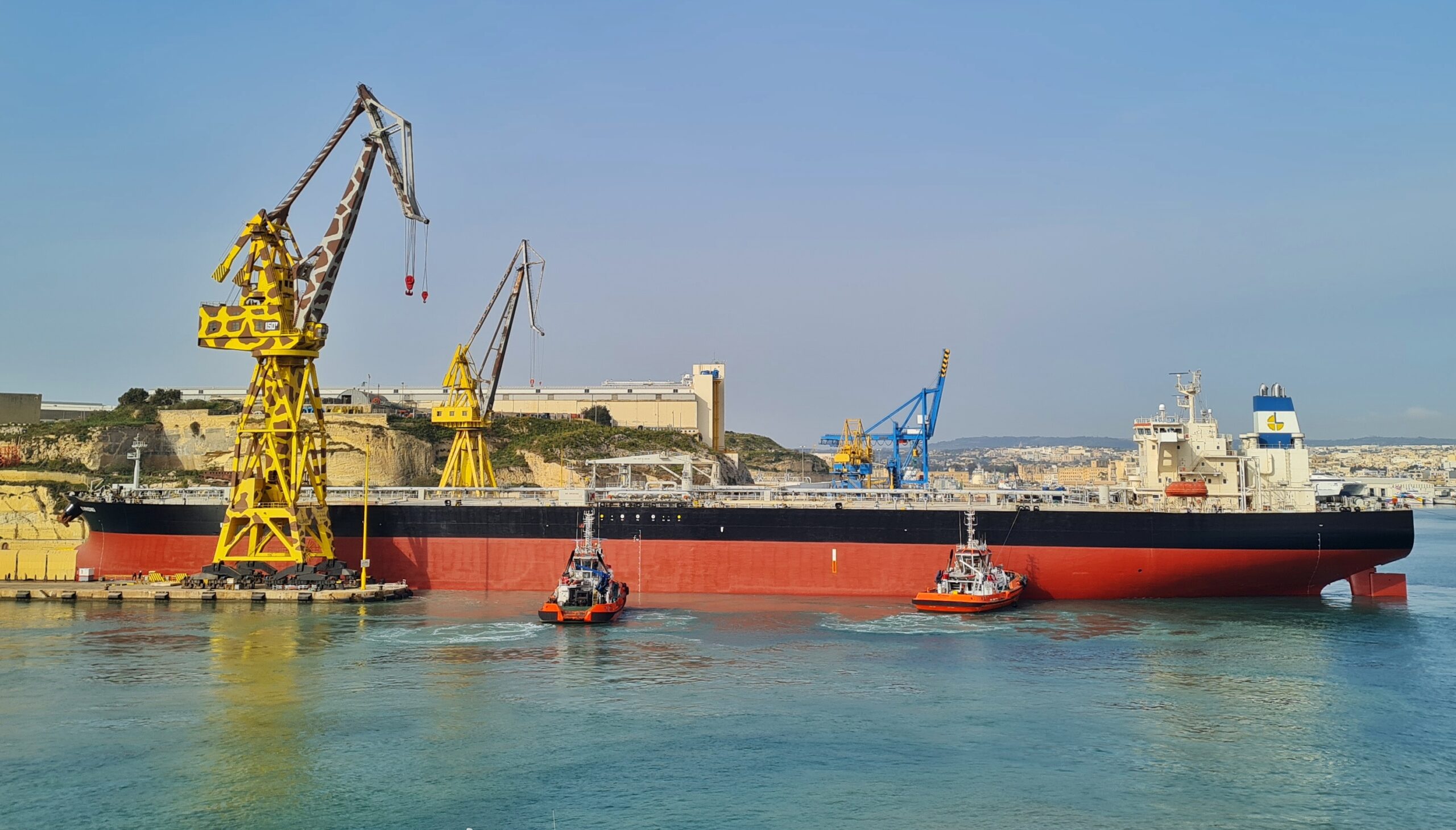 Ein großes und sehr langes Schiff (der untere Rumpf rot; darüber ein breiter schwarzer Streifen) fährt rückwärts aus einem Dock. Im hinteren Bereich befindet sich ein hoher weißer Aufbau mit dem Steuerstand; ansonsten hat das Schiff keine Aufbauten). Zwei kleine, rote Schlepper fahren seitlich (das heißt aus Richtung der Kamera) auf das Schiff zu. Links sind (vor und hinter dem Schiff) zwei große Kräne zu sehen. Einer gelb mit braunen Flecken, der andere gelb. Im Hintergrund ein weiterer blauer Kran sowie Gebäude.
