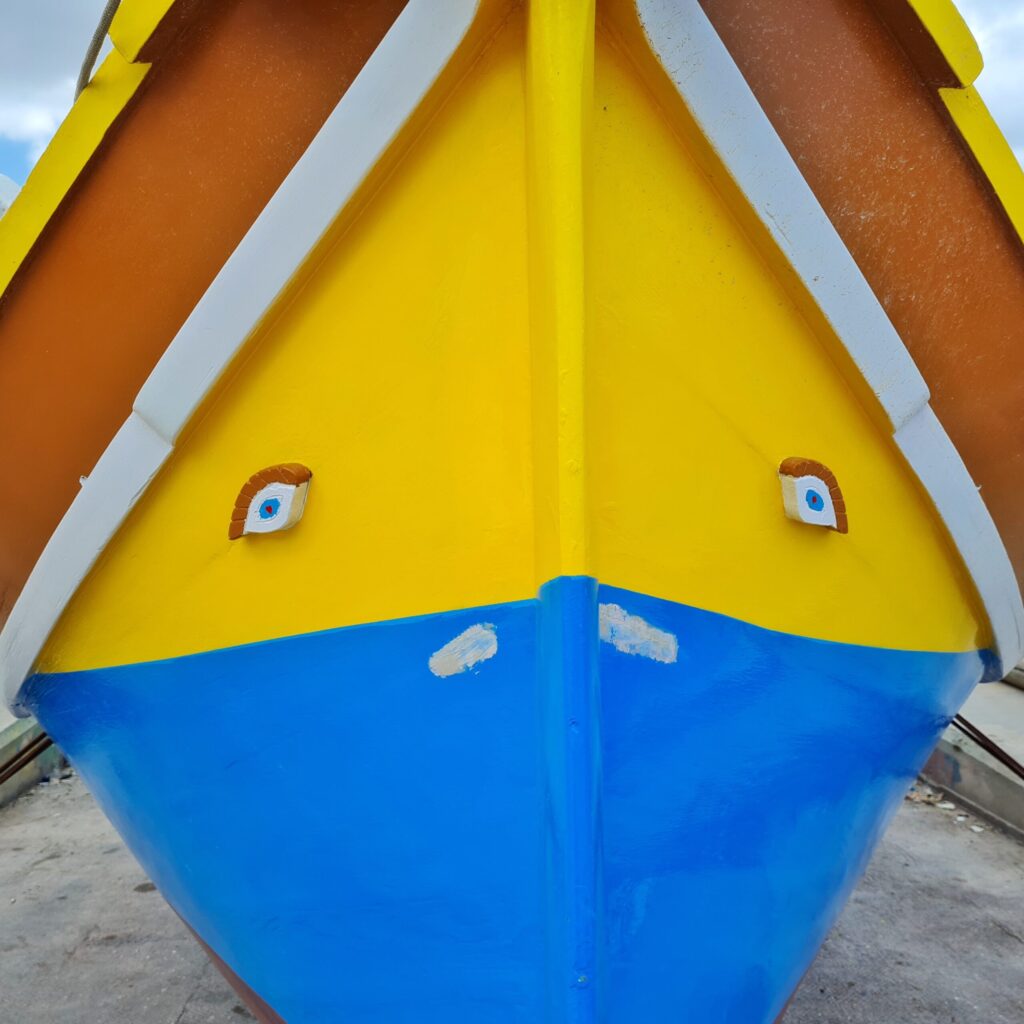 Ein Boot, das an Land liegt, in Großaufnahme. Unten ist es hellblau. In dem gelben Teil darüber sind links und rechts blaue Augen mit braunen Wimoern angebracht. Der obere Teil des Bootes, der auf dem Foto ein Dreieck bildet, besteht aus einem breiten bräunlichen Streifen, darüber ein schmaler gelber und darunter ein schmaler weißer. 